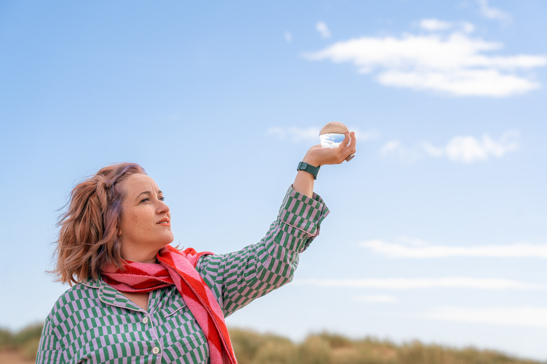 A white woman with ginger and lilac hair and freckles raises a crystal ball to the blue sky, looking directly at it.