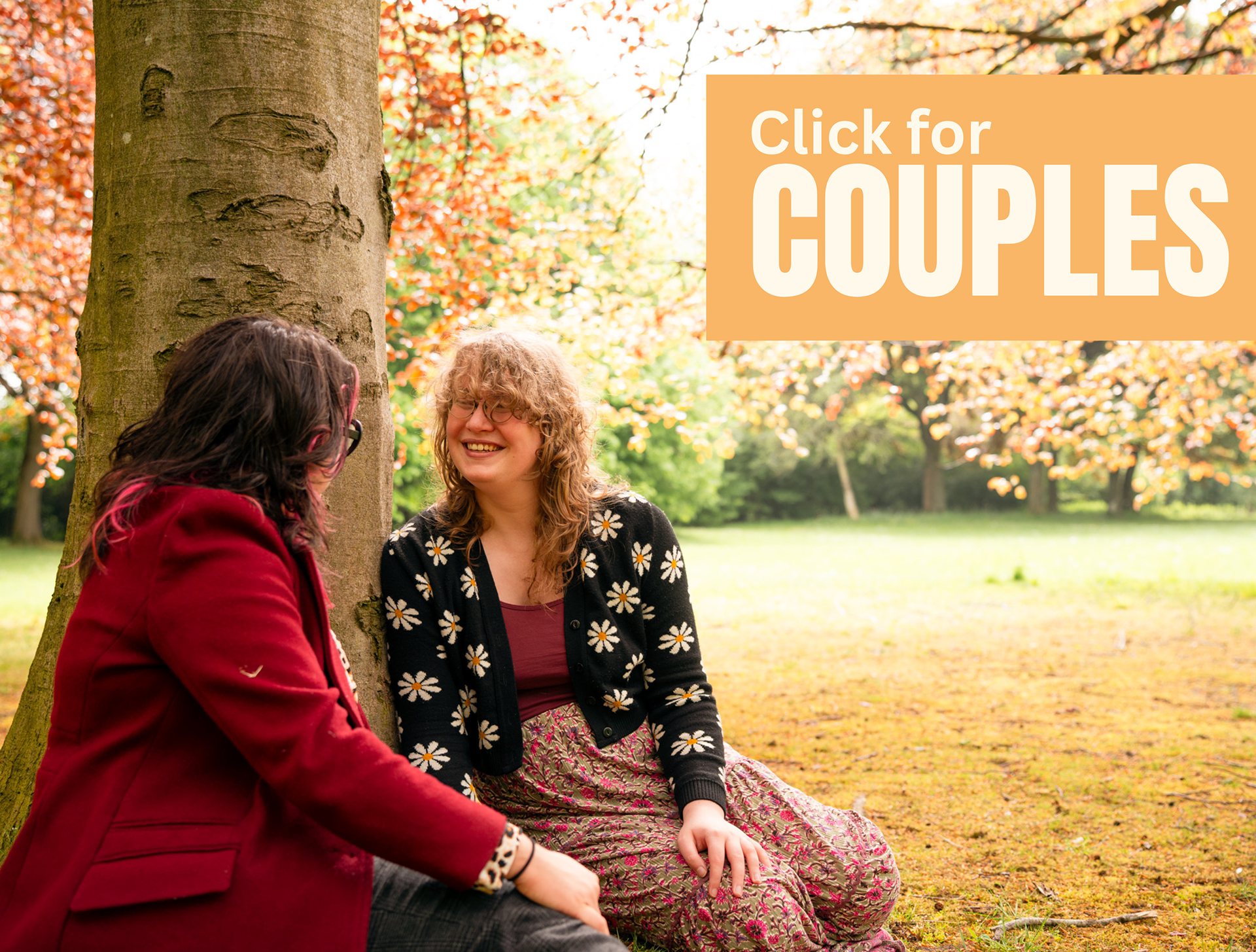 In an autumnal park, a young couple, both with long wavy hair, sit under a tree together and smile at one another. He has pink streaks through his dark hair and a red coat; she wears a floral cardigan and long skirt. The overlaid text reads "Click for Couples".