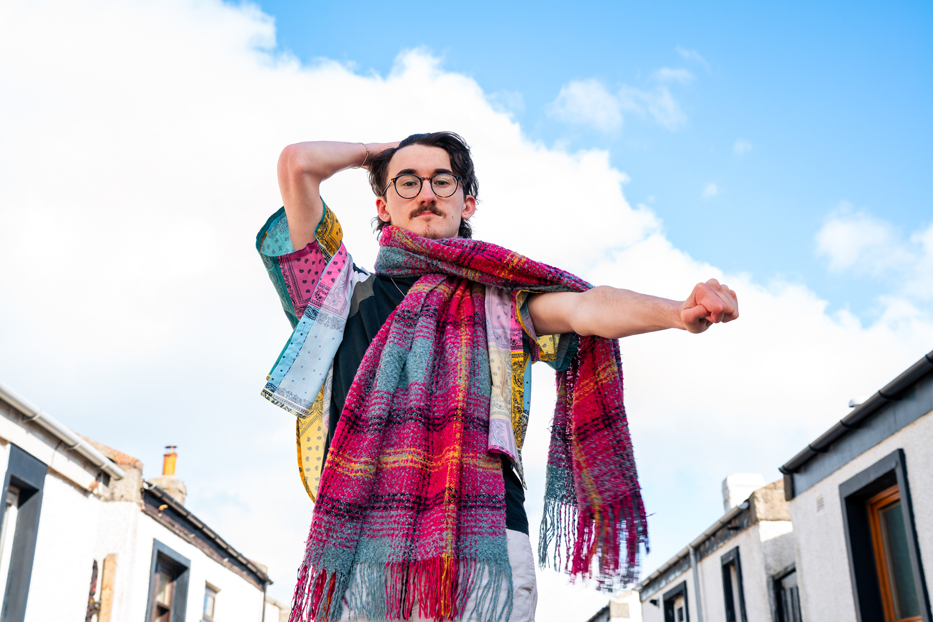 A white man with cerebral palsy is seen from below, towering over the camera, one arm help forward proudly, the other pushing his hair out of his face. Blue sky and white clouds behind him.