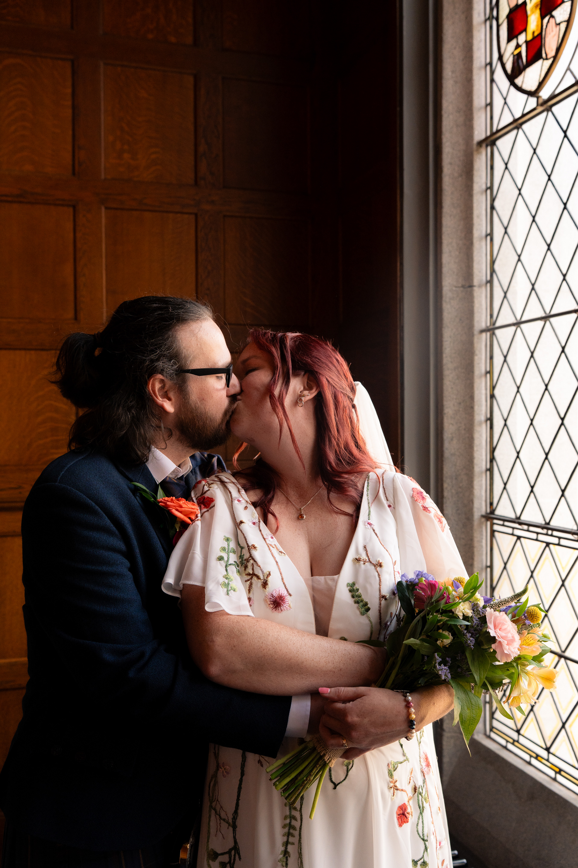 A long-haired groom in navy and red-haired bride in floral dress kiss, lit by the light from a stained glass window.