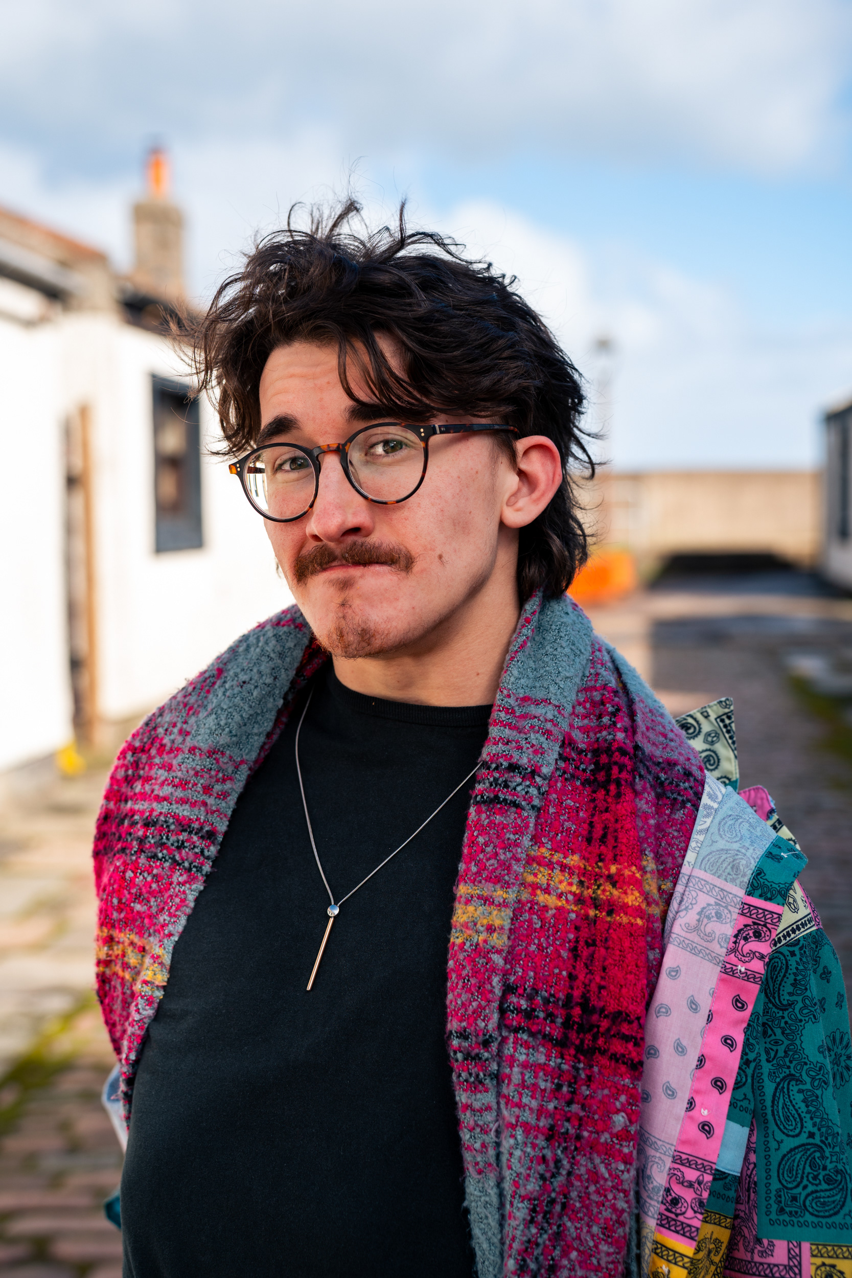 A handsome man with messy hair, small beard and glasses looks directly at the camera. Fittie, Aberdeen is the backdrop.