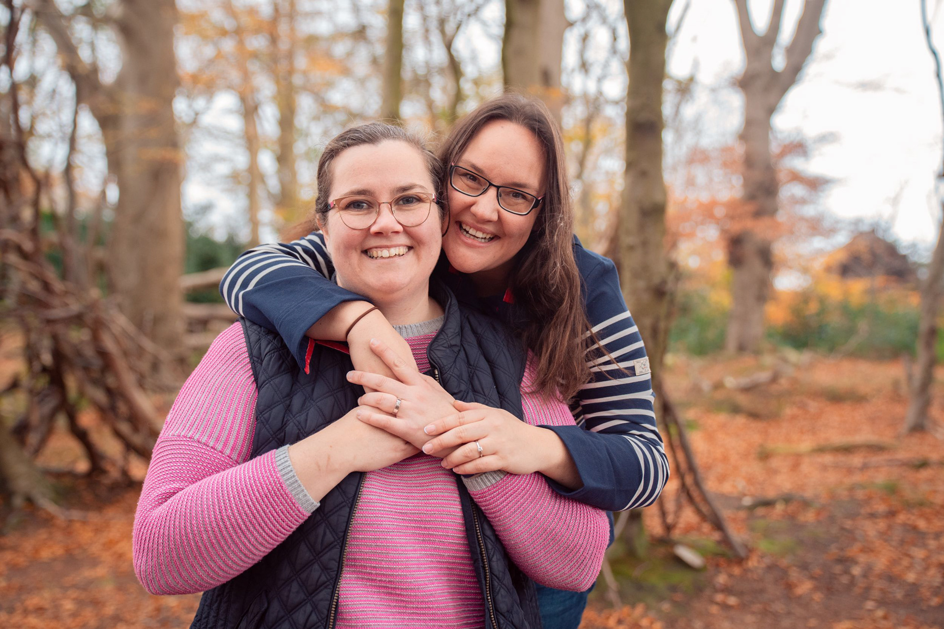 Two brunette women in striped tops stand with their arms around each other and engagement rings showing, in autumn woodland, on their engagement photo session.