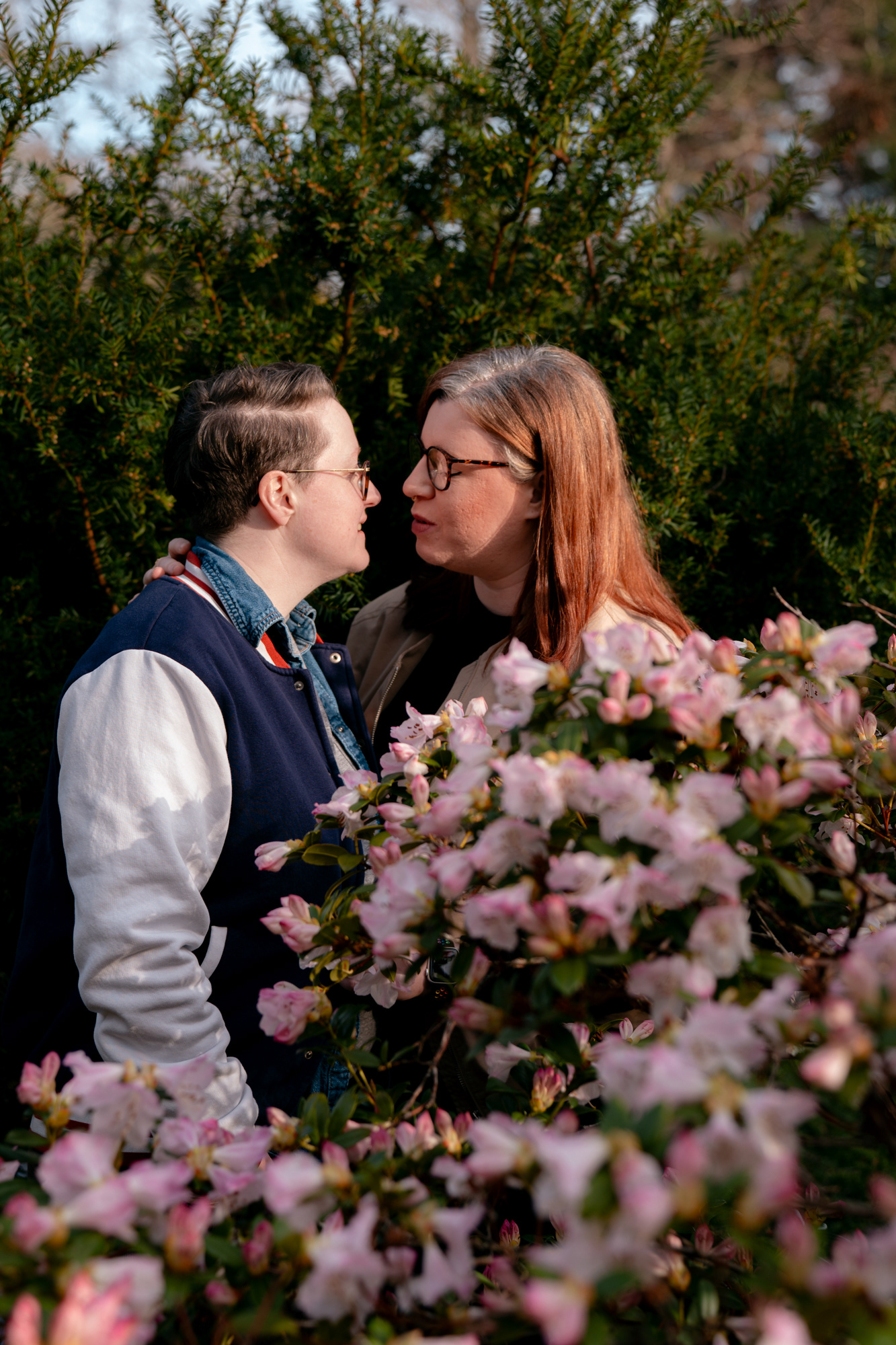 A queer couple (she/her and they/them) come in for a kiss, surrounded by pink blossom.