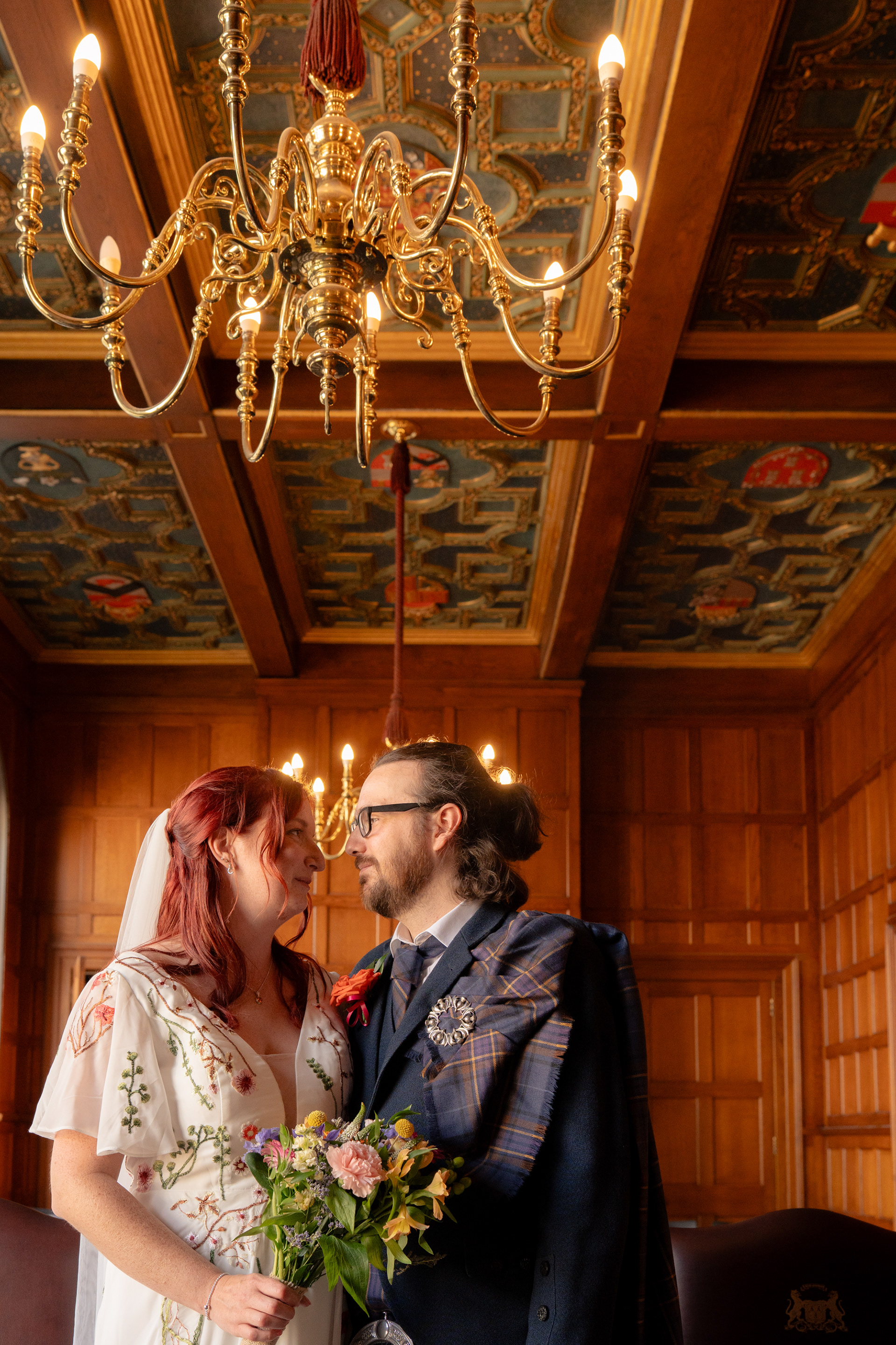 The ornate ceiling and chandeliers in Marischal College's Grant Room are visible behind this bride in floral dress and groom in blue kilt as they turn to face each other.