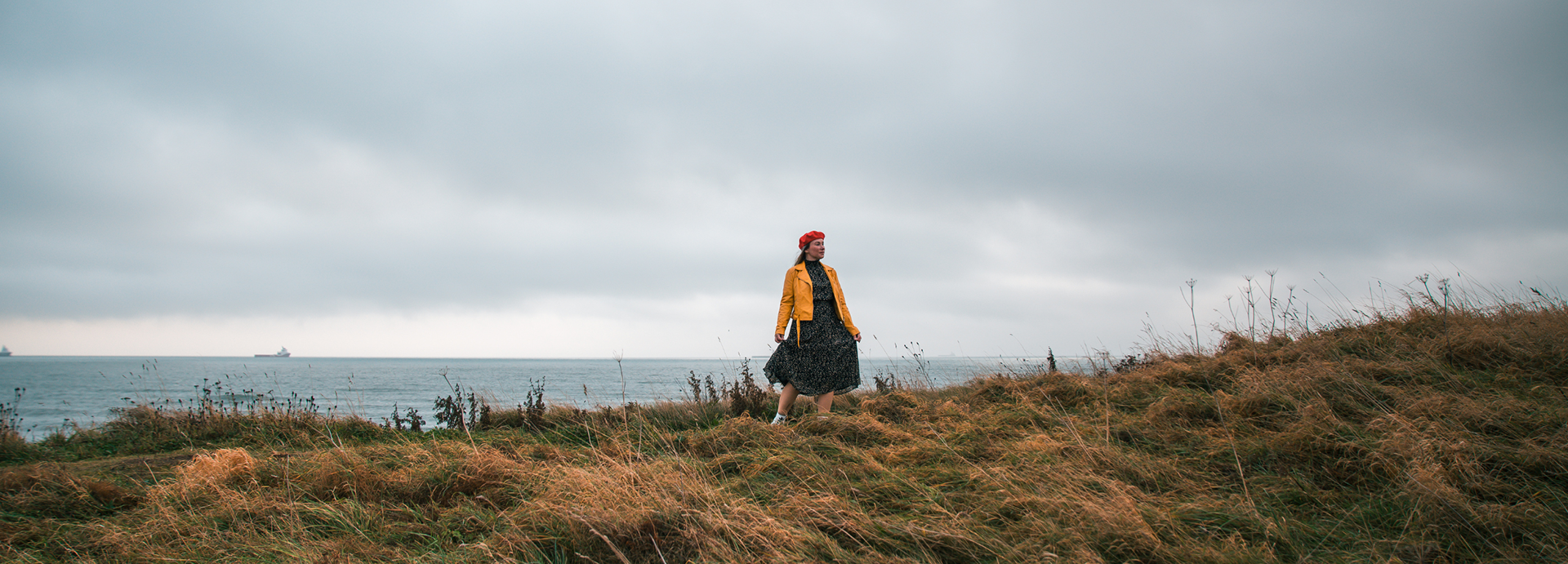 On an Aberdeen main character empowerment shoot, a woman with green hair, wearing a black dress, yellow coat and red beret, stands on a grassy dune with the North Sea behind her.