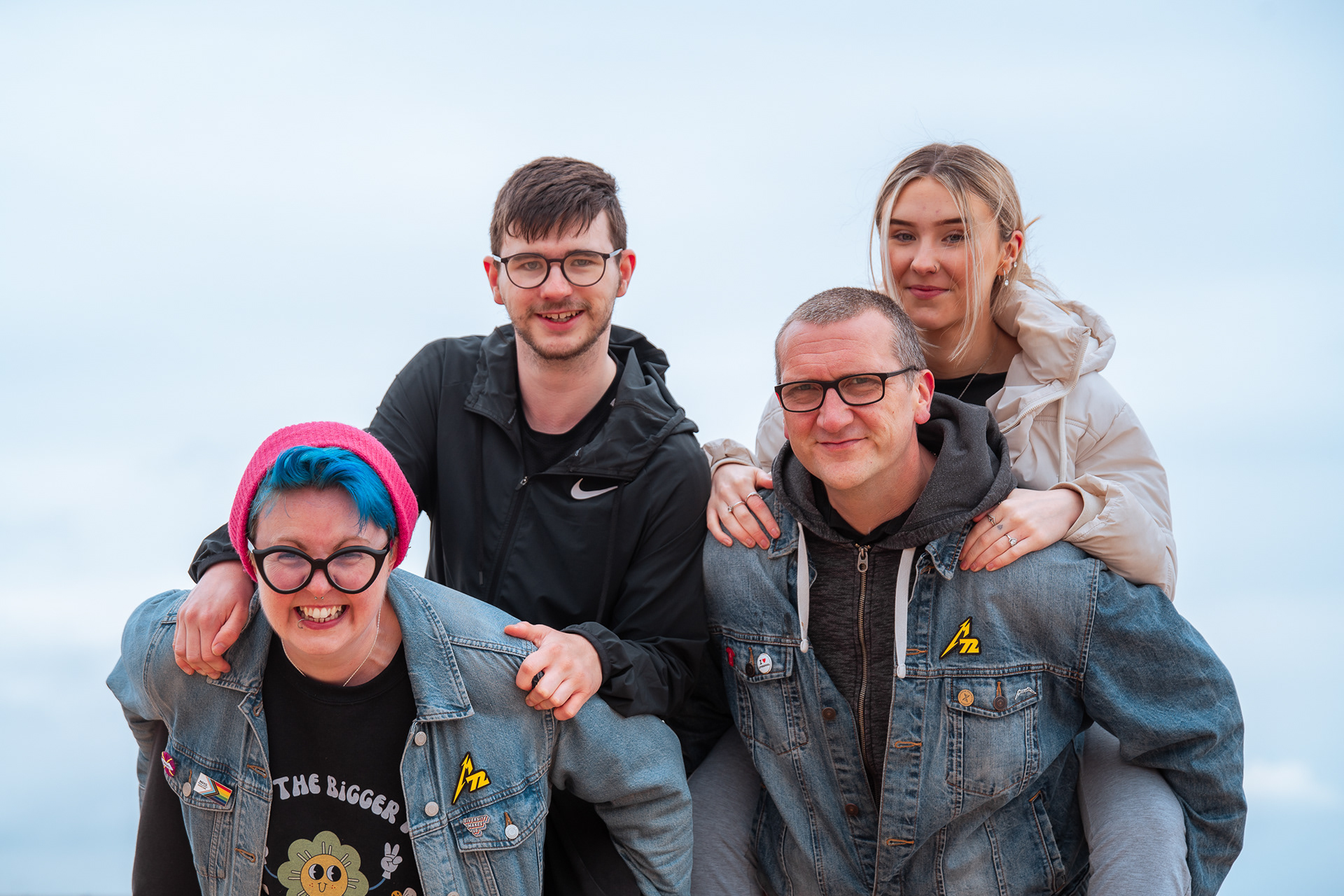 Two grown ups in denim jackets - the woman with blue hair and piercings - give piggybacks to teenagers; all four smile directly at the camera