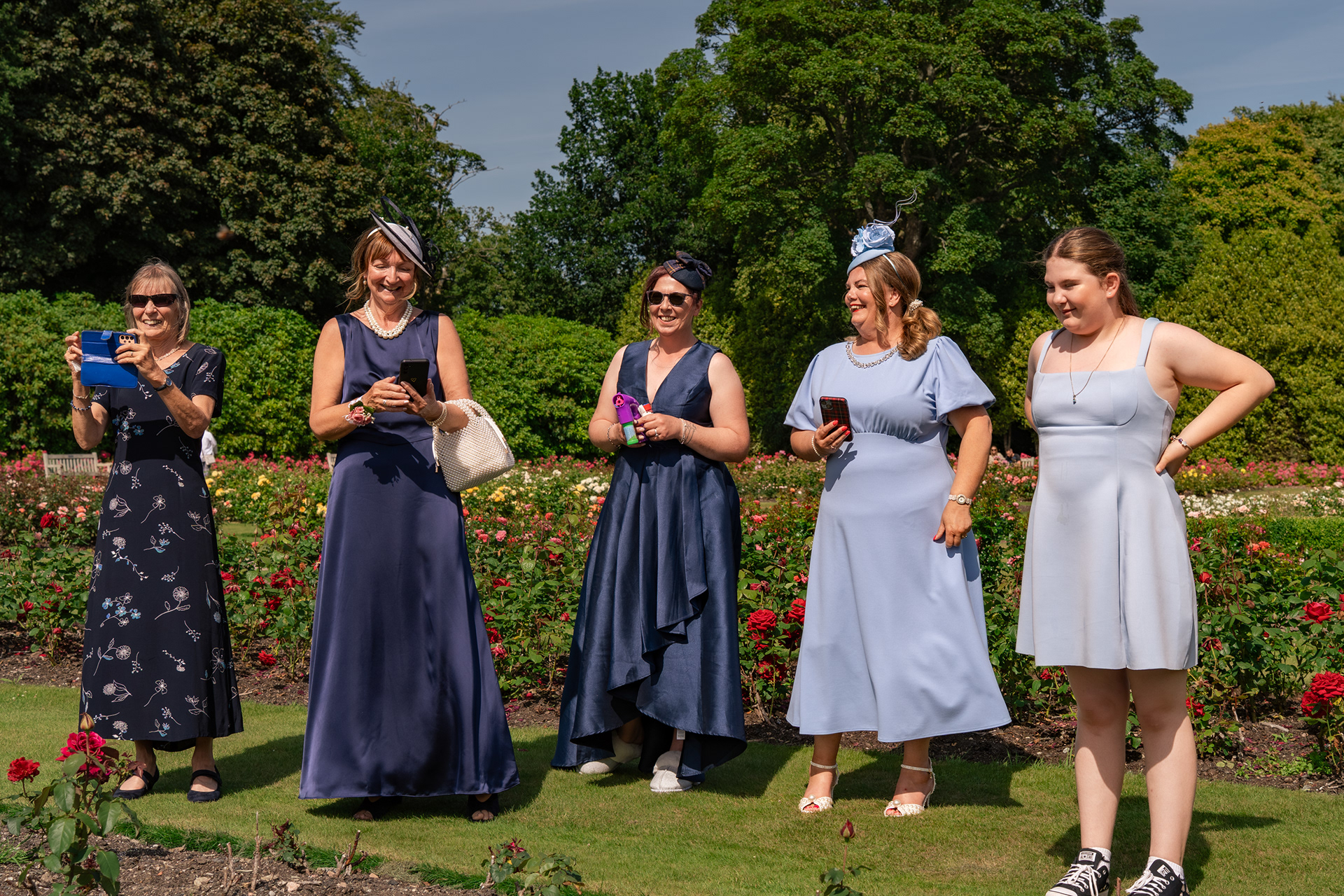 Five women in assorted shades of blue stand, smiling and laughing, in the rose gardens at Hazlehead, watching group wedding photos being taken