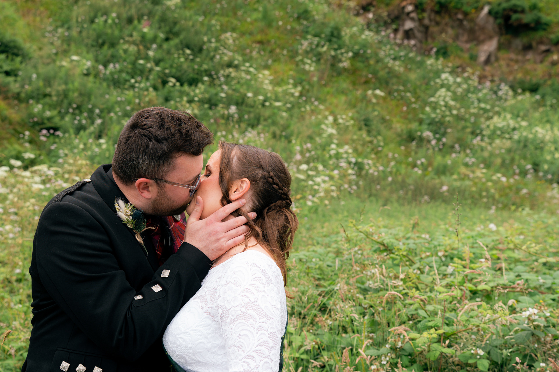 Stonehaven registry office wedding photography: bride in white and groom in kilt kiss against a backdrop of greenery.
