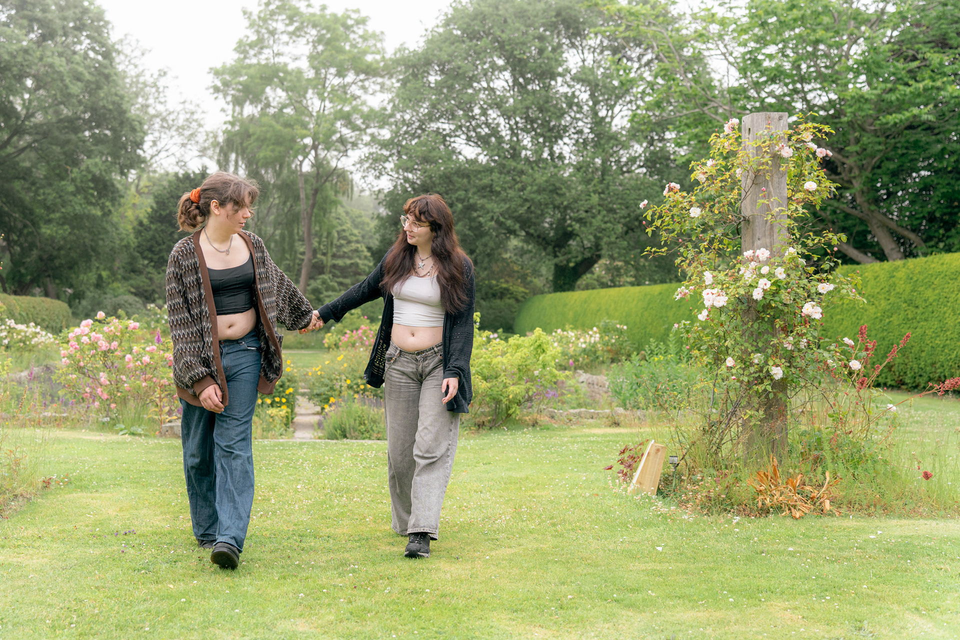 A young couple in jeans and long cardigans, hold hands and swing their arms as they stroll across a misty garden in Aberdeen.