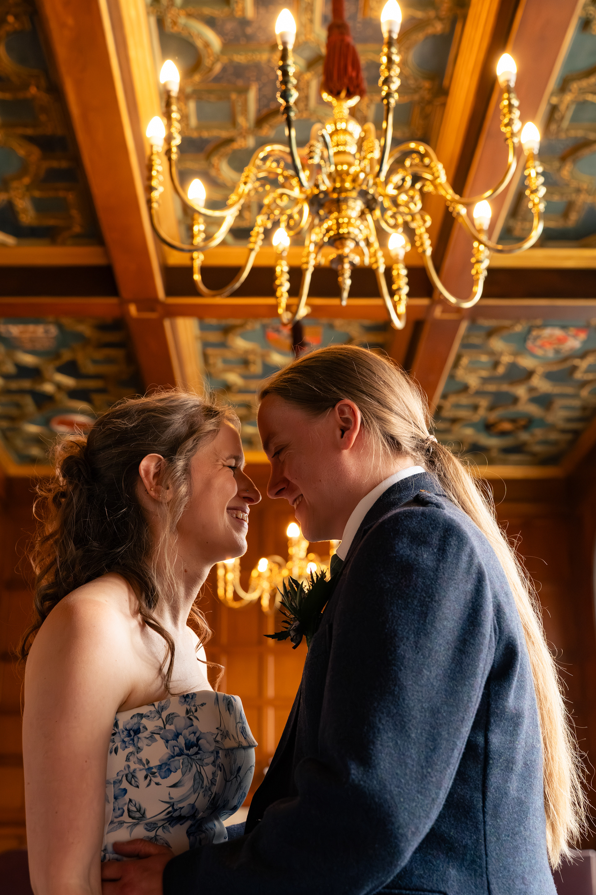 Aberdeen Wedding: A newlywed couple (she/her and he/him), both with long brown hair tied back, grin at each other under the ornate blue and gold ceiling and chandelier in the Marischal College registry office in Aberdeen.