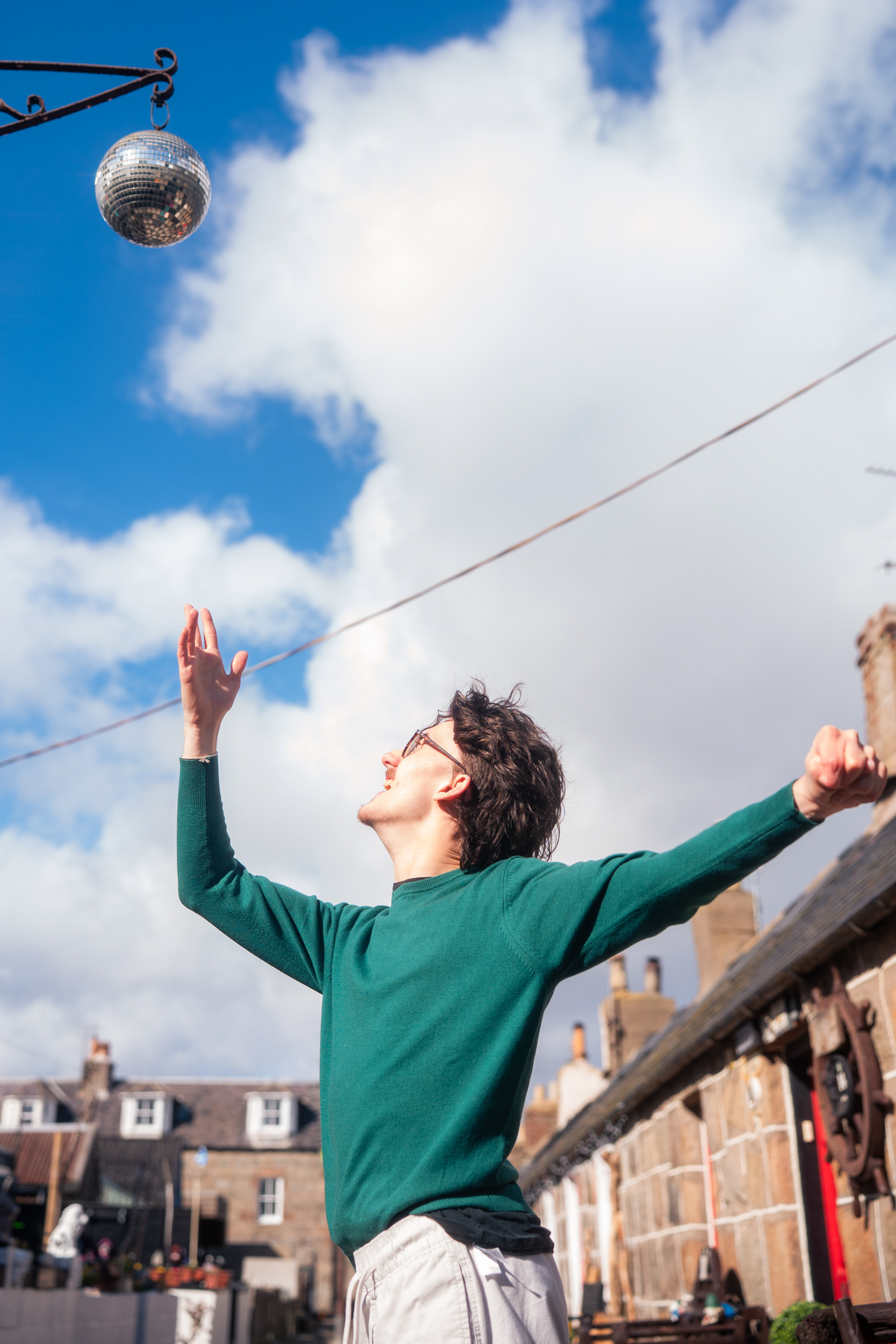 A Disabled man in a green jumper strikes a pose beneath a disco ball and a blue sky in Fittie, Aberdeen.