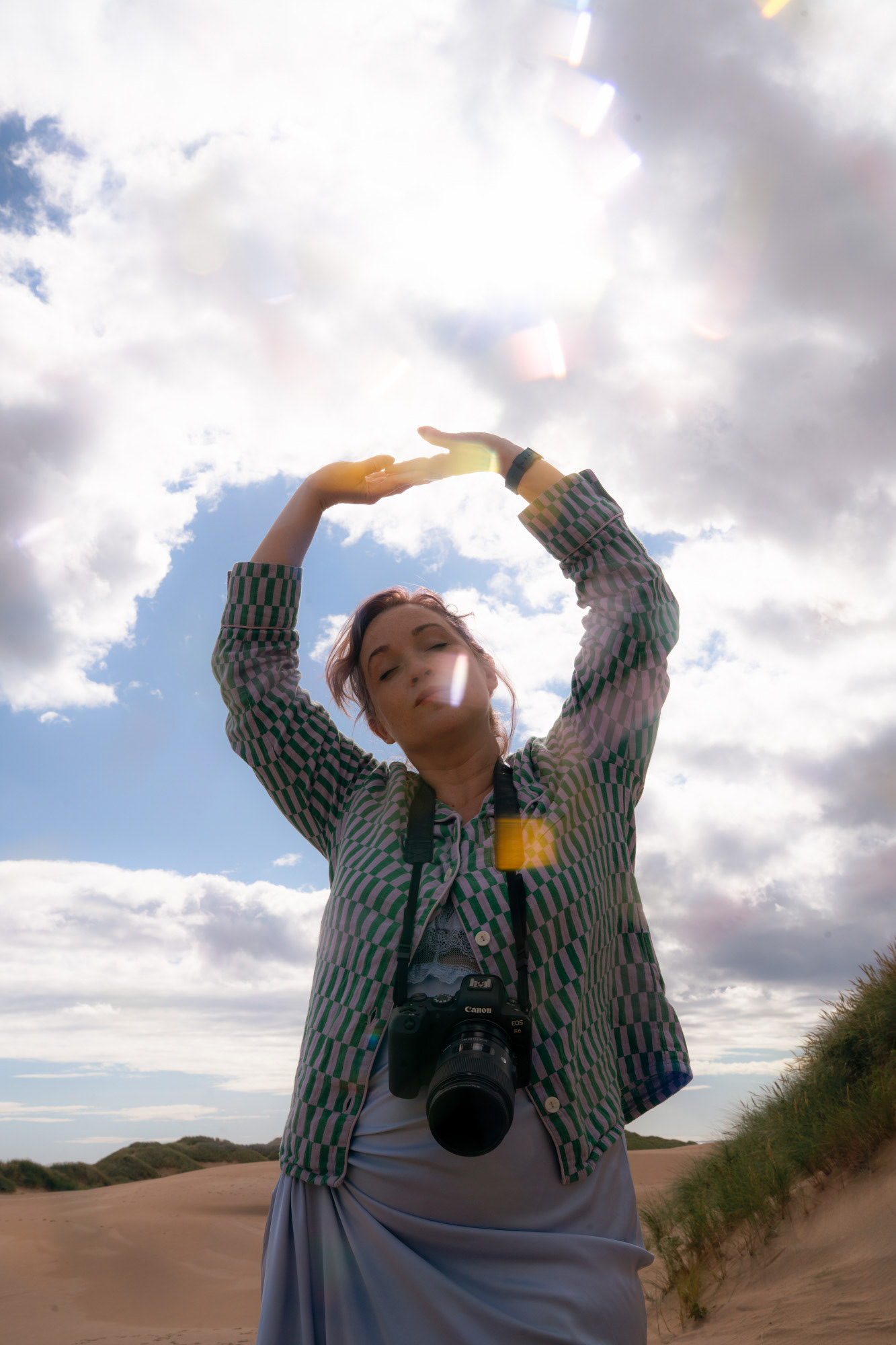 A white woman with a ponytail, pyjama top and a camera hanging around her neck, raises her hands to a cloudy sky, with sun flare speckling over her.