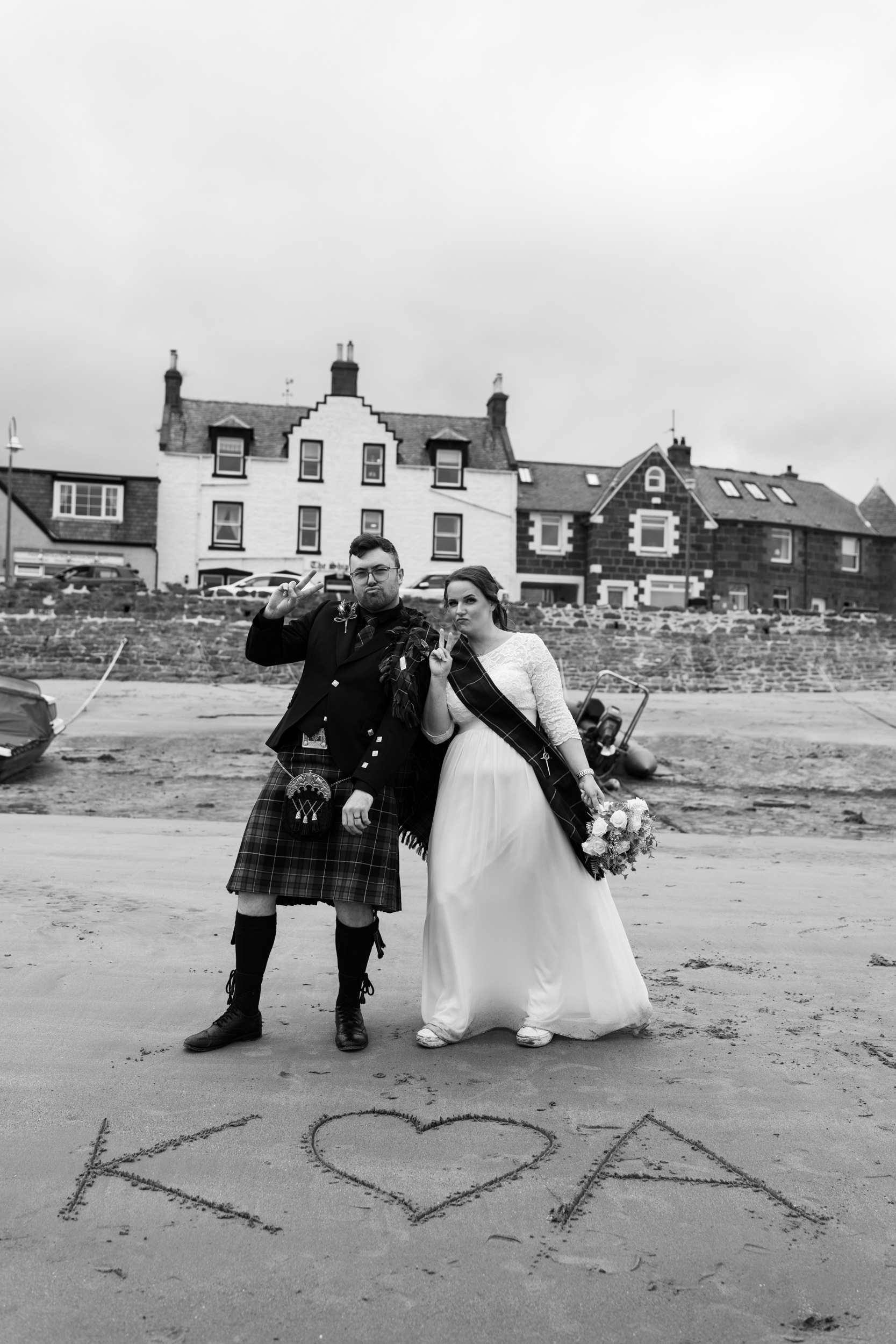Alternative Aberdeenshire wedding photography: Bride and groom make peace signs, their initials written in the sand in front of them, following their Stonehaven registry office elopement.