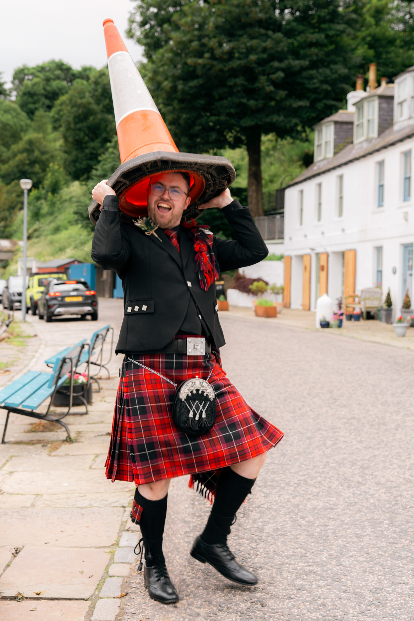 Alternative Aberdeenshire wedding photography: A groom in red kilt lifts a traffic cone over his head like a hat.