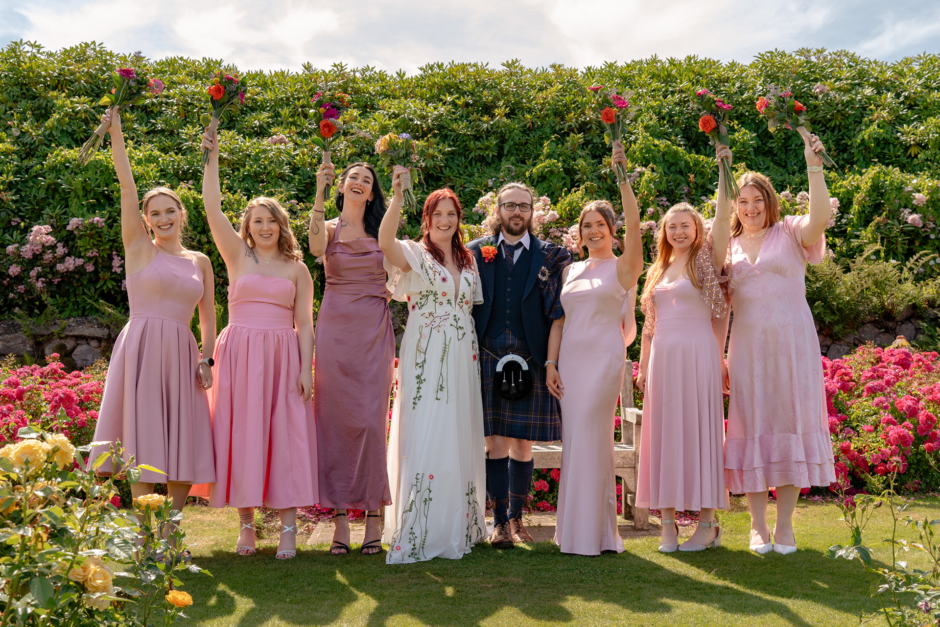 Bride in floral dress and groom in blue kilt stand with six bridesmaids in different pale pink dresses. The women all raise their bouquets above their heads. They are surrounded by magenta rose bushes in the Hazlehead Park gardens.