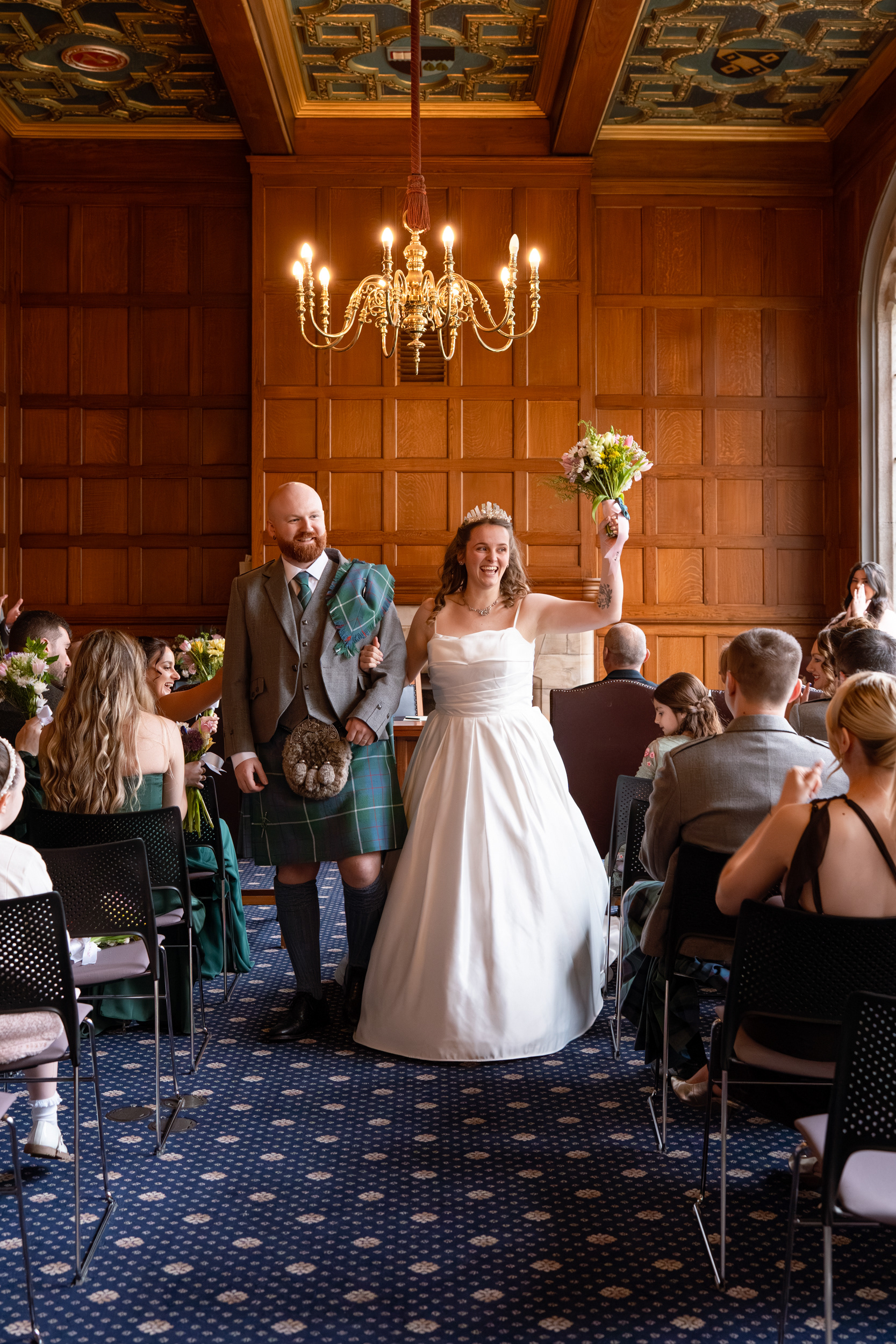 Newlywed couple walk back down the aisle in the Marischal College Grant Room, bride's bouquet raised in celebration, as their guests cheer them on.