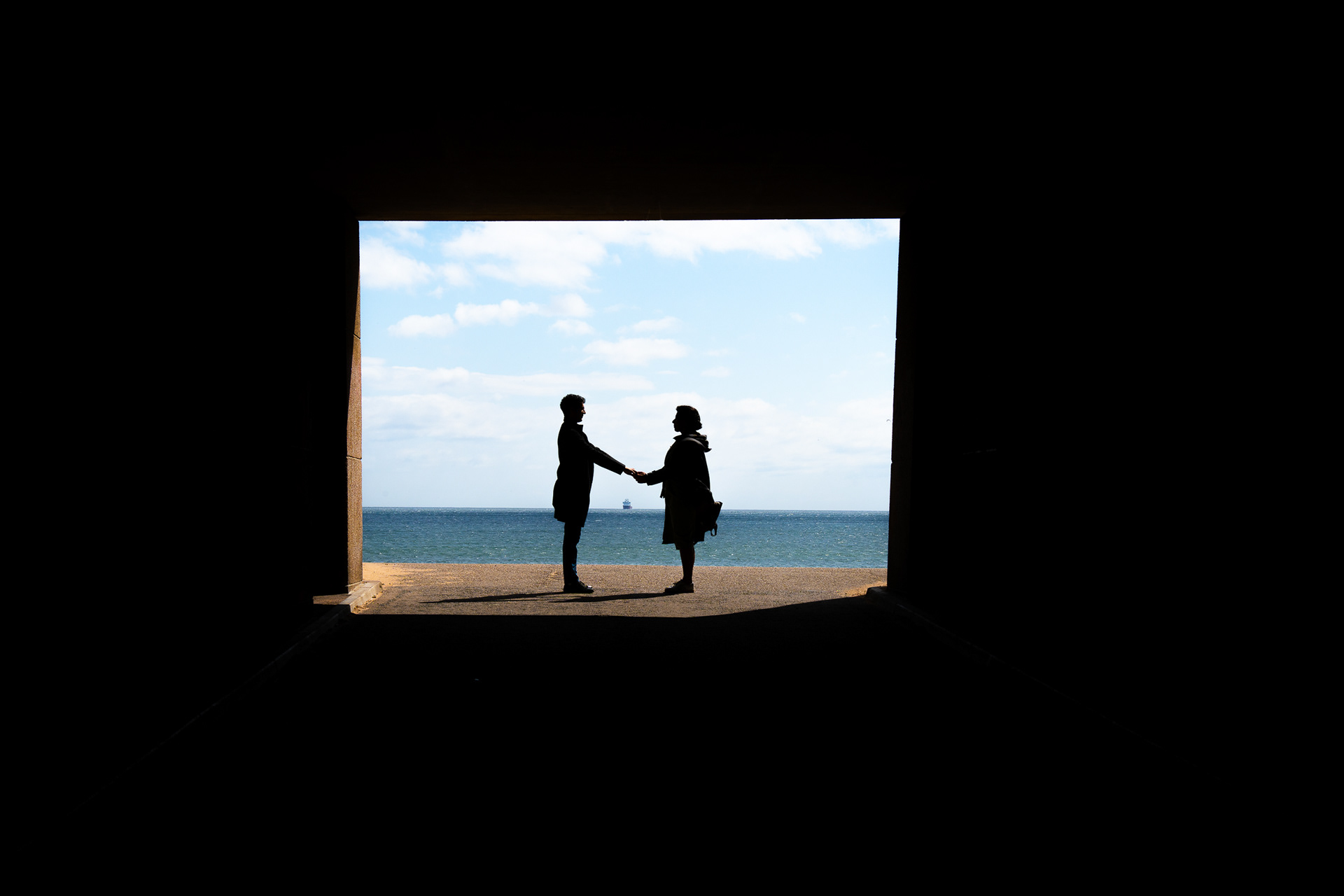 A silhouetted couple stand face to face, holding hands, at the entrance to a pitch black tunnel. The North Sea is visible behind them; the sky and sea are bright blue.