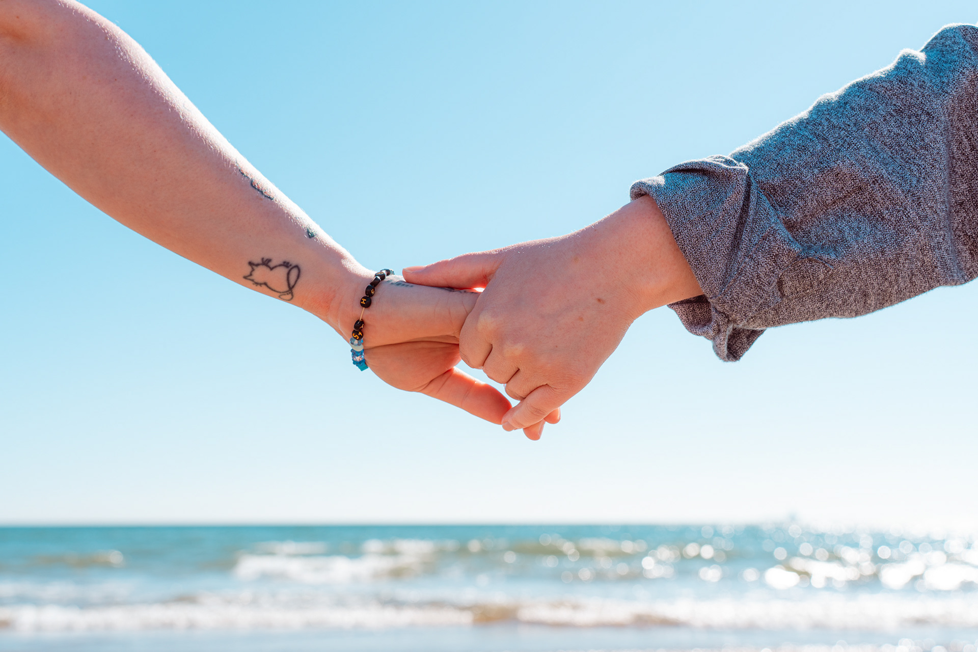 Hands clasped against a backdrop of blue skies and sparkling sea. One hand has a cat tattoo on the wrist and she wears a Trans Pride bracelet. The other hand belongs to her non-binary partner, grey shirt sleeve showing.