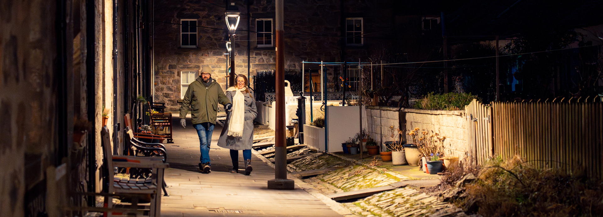 A male-female couple in winter coats stroll through Fittie (Footdee) after dark, lit by lamplight, during their Aberdeen couple's photo shoot