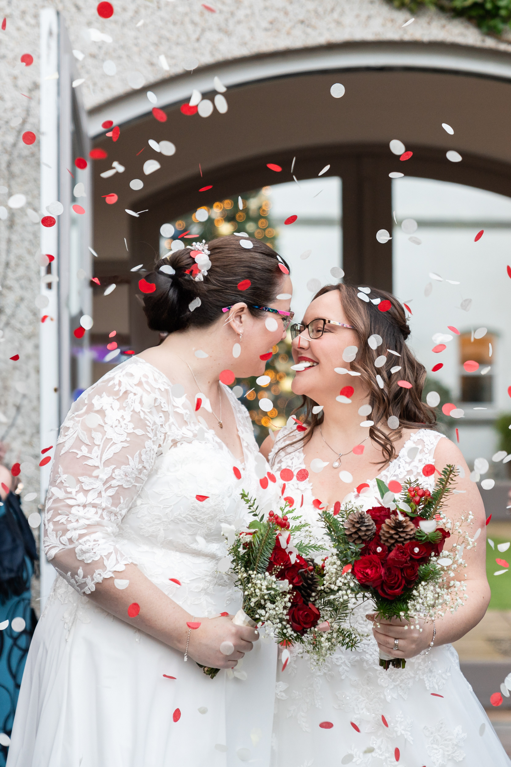 Two brides in white dresses lean in to kiss as red and white confetti drifts down around them.