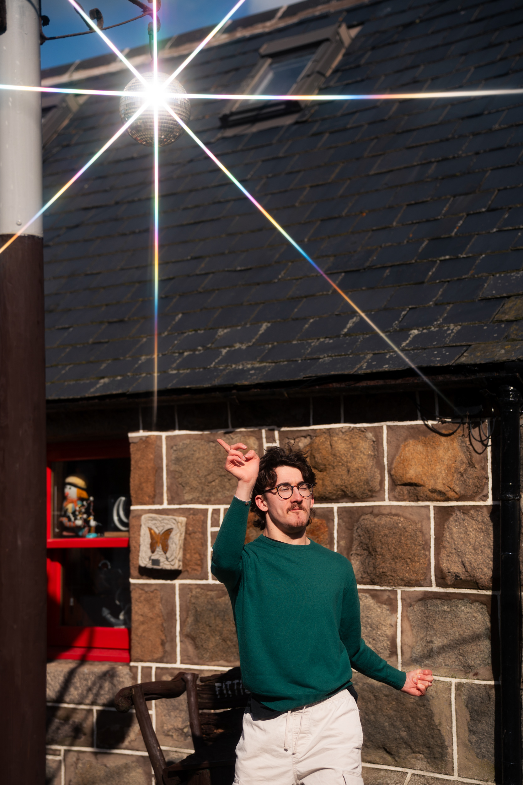 A young man in green points his finger and disco dances as light bounces off the glitter ball hanging above him.