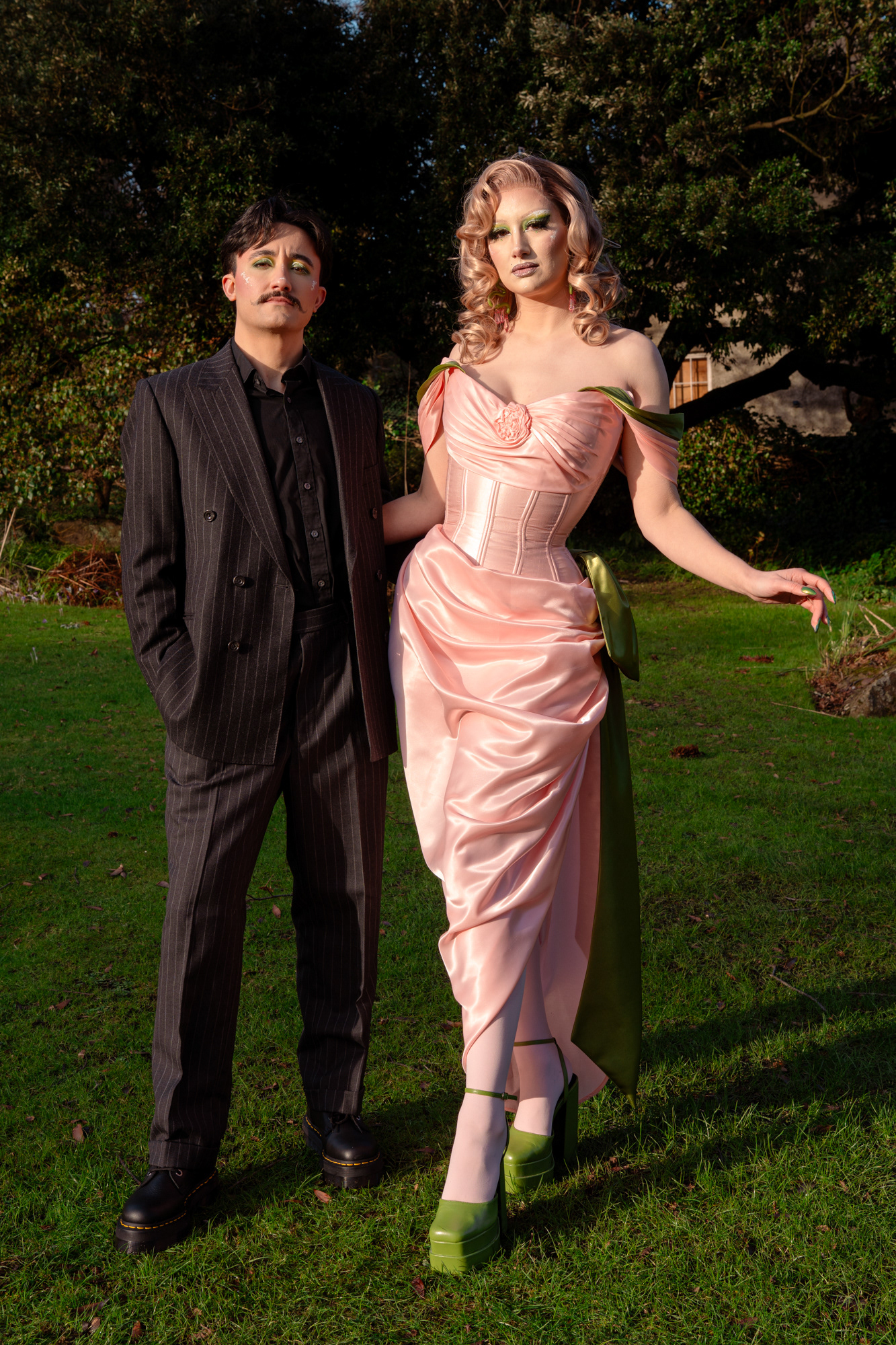 A couple in drag pose together during their Aberdeen couple's photo session. They/them is wearing a pinstripe suit and has short, dark hair and a moustache. She/her is in a glamorous pink and green dress and high green heels with dramatic makeup. They are standing next to each other, looking straight at the camera.