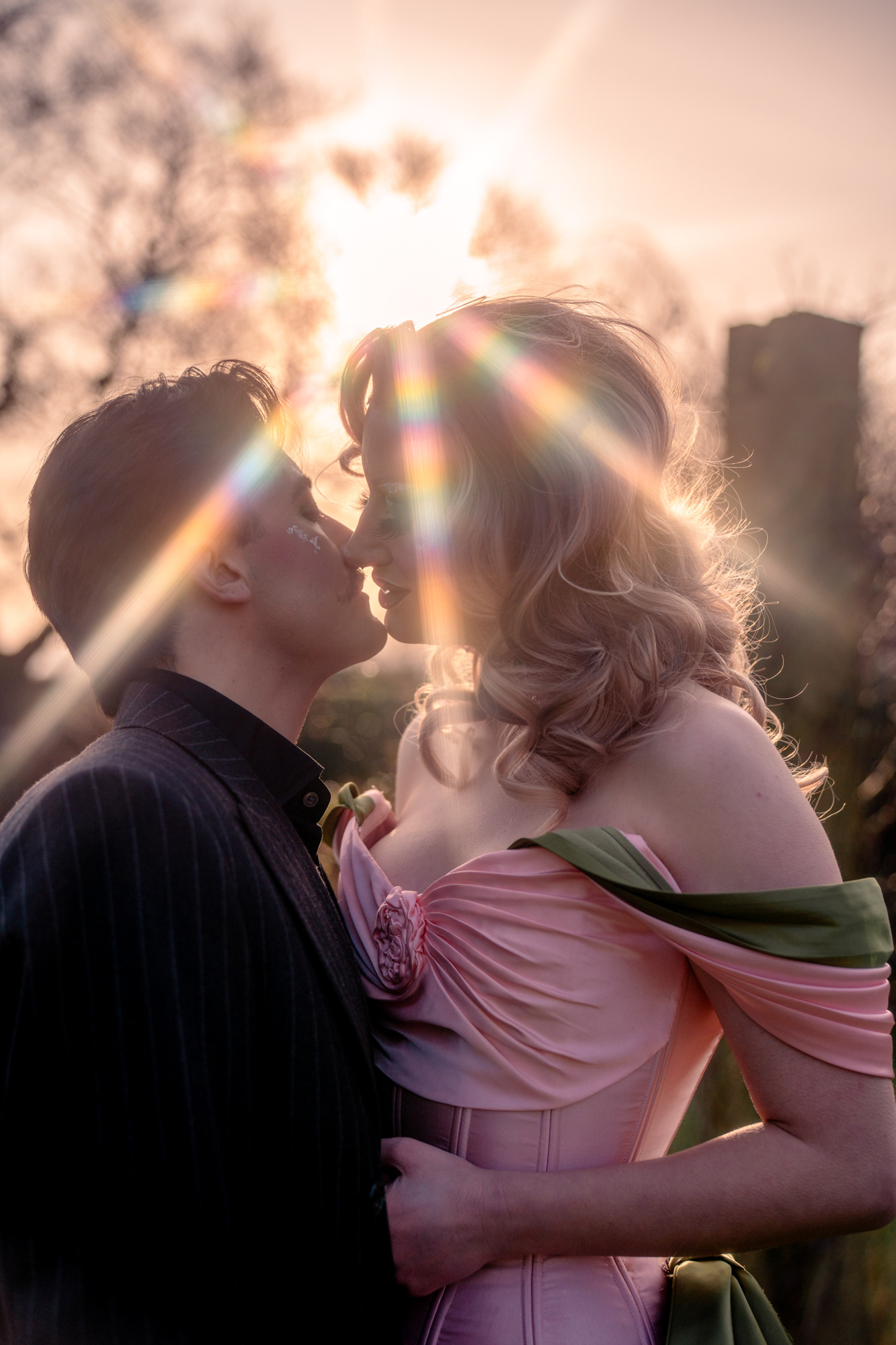A couple in drag pose together during their Aberdeen couple's photo session. They/them is wearing a pinstripe suit and has short, dark hair and a moustache. She/her is in a glamorous pink and green dress and high green heels with dramatic makeup. They are leaning in for a kiss with a rainbow starburst of light above them.