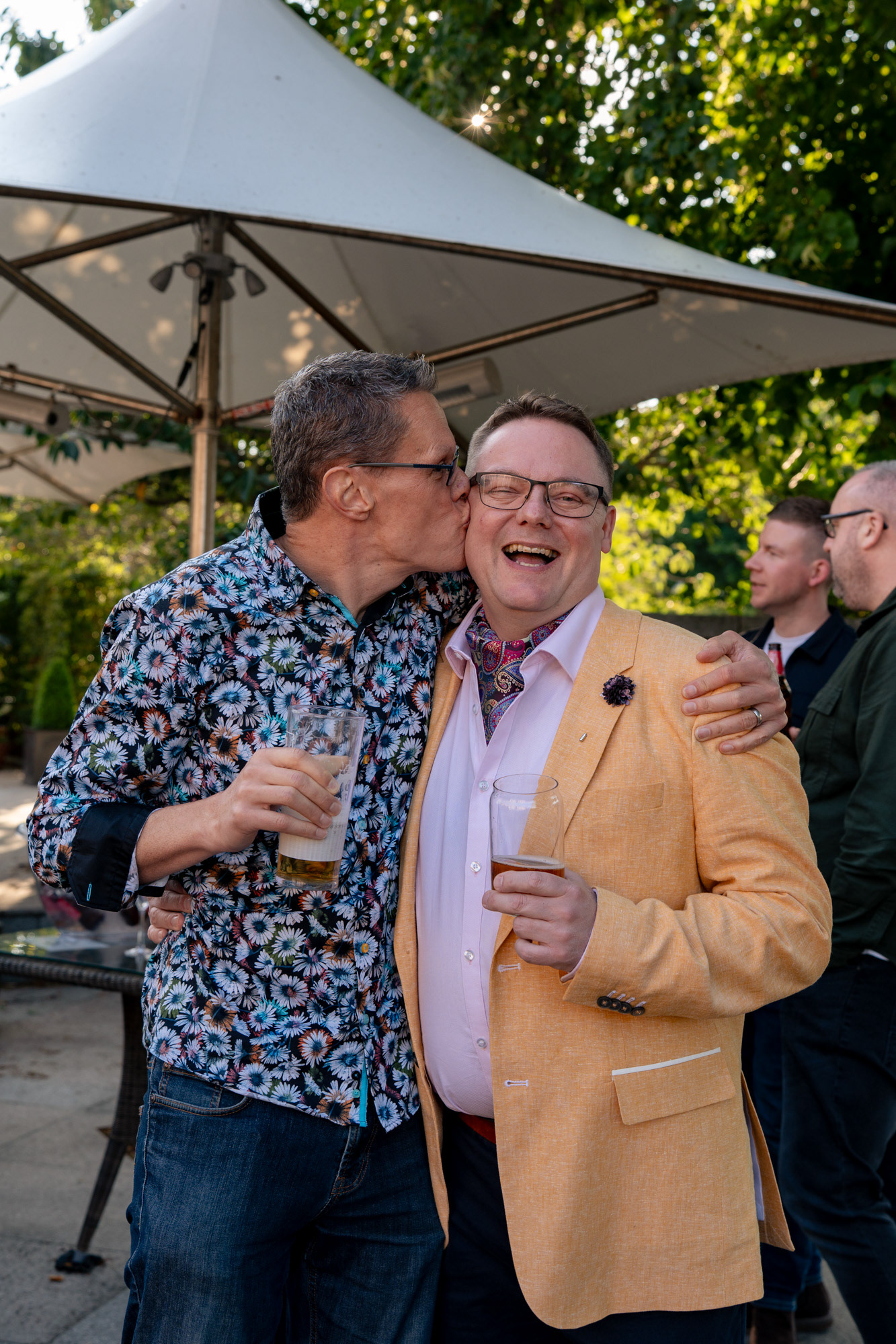 Two men celebrating fiftieth birthdays, beers in hands. One, in a floral shirt, kisses the other, in a yellow jacket and cravat, on the cheek as his friend laughs and looks directly at the camera.