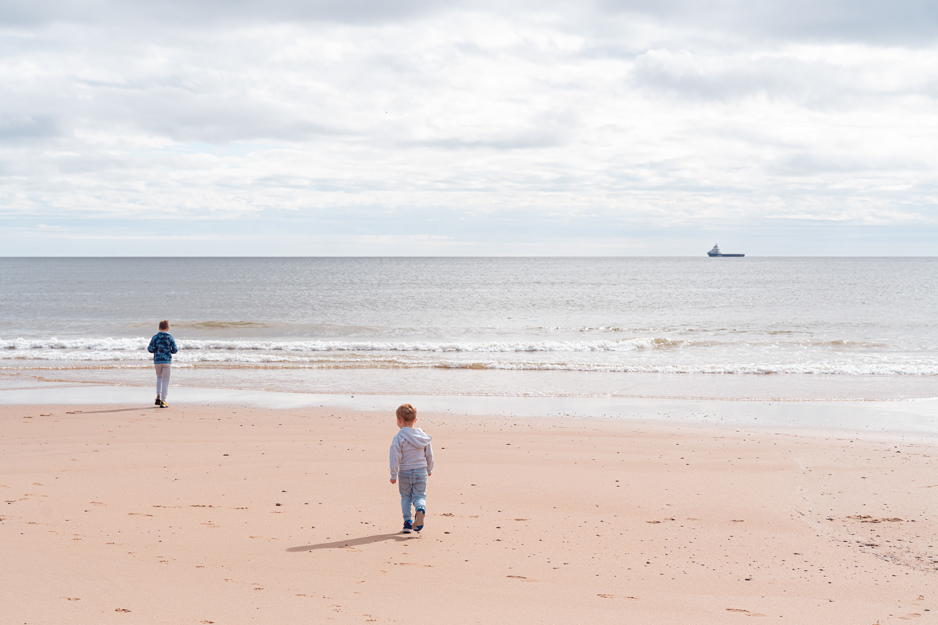Two young boys in blue at an Aberdeen family photo shoot walk across the sand towards the shining blue sea at Aberdeen Beach.