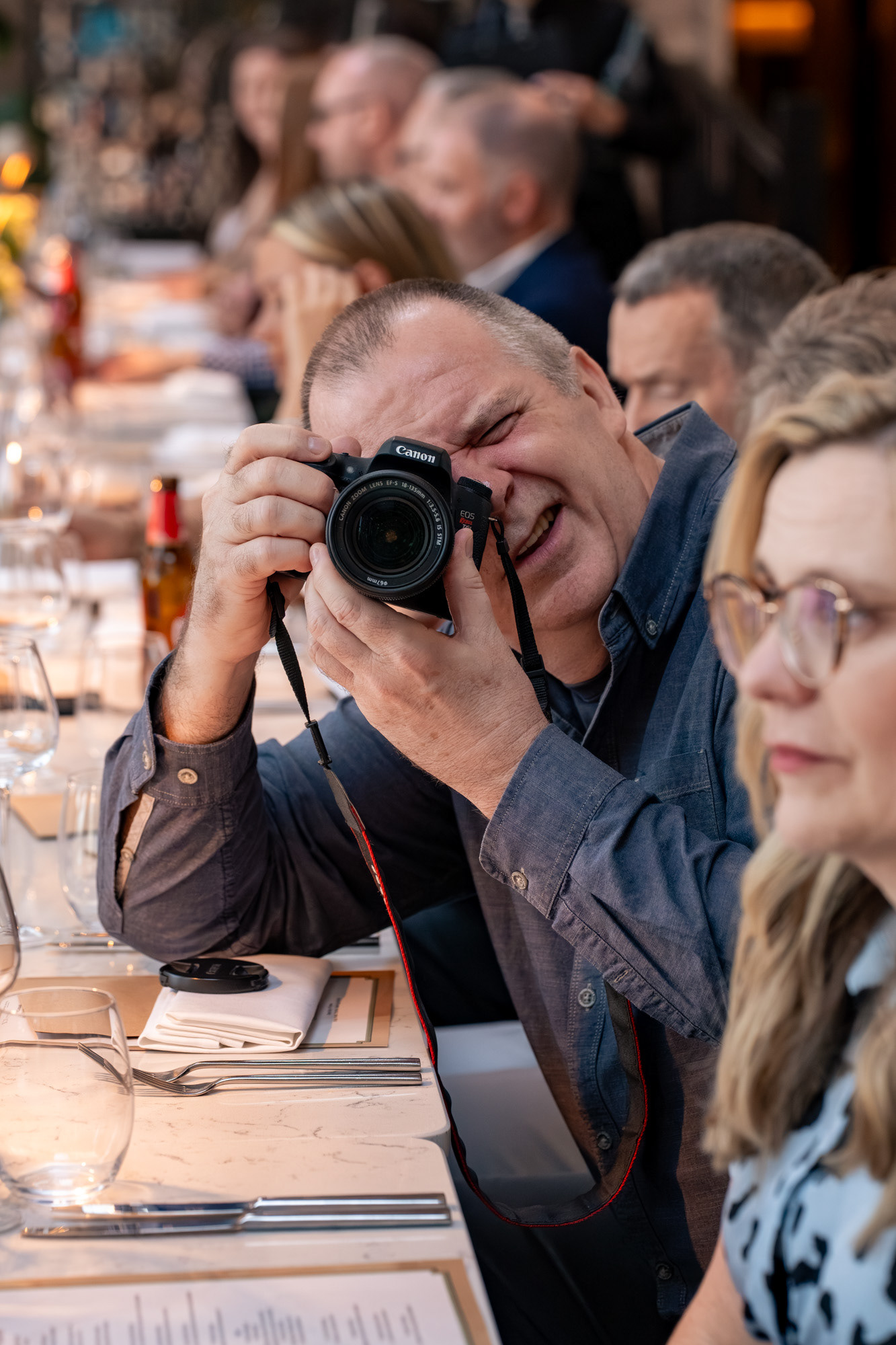 A man in a grey shirt points his Canon camera directly at the photographer.