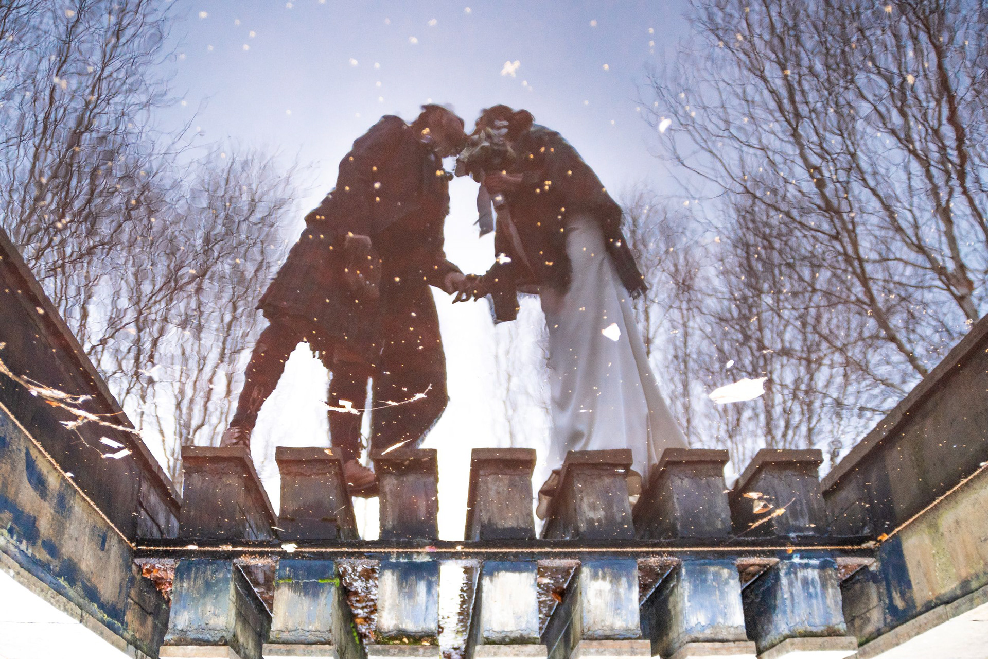 Aberdeen Wedding Photography: A newlywed couple, he in a kilt and she in a white dress and tartan shawl, are reflected in a pond as they kiss. The trees behind them are wintry.