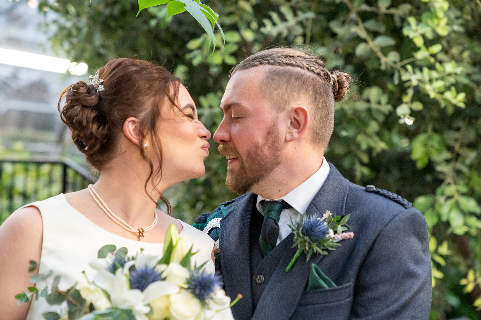 Duthie Park Wedding Photography: A bride in ivory puckers up in a comedy fashion as she leans towards her new husband, dressed in navy and green kilt attire with plaits holding back his long hair.
