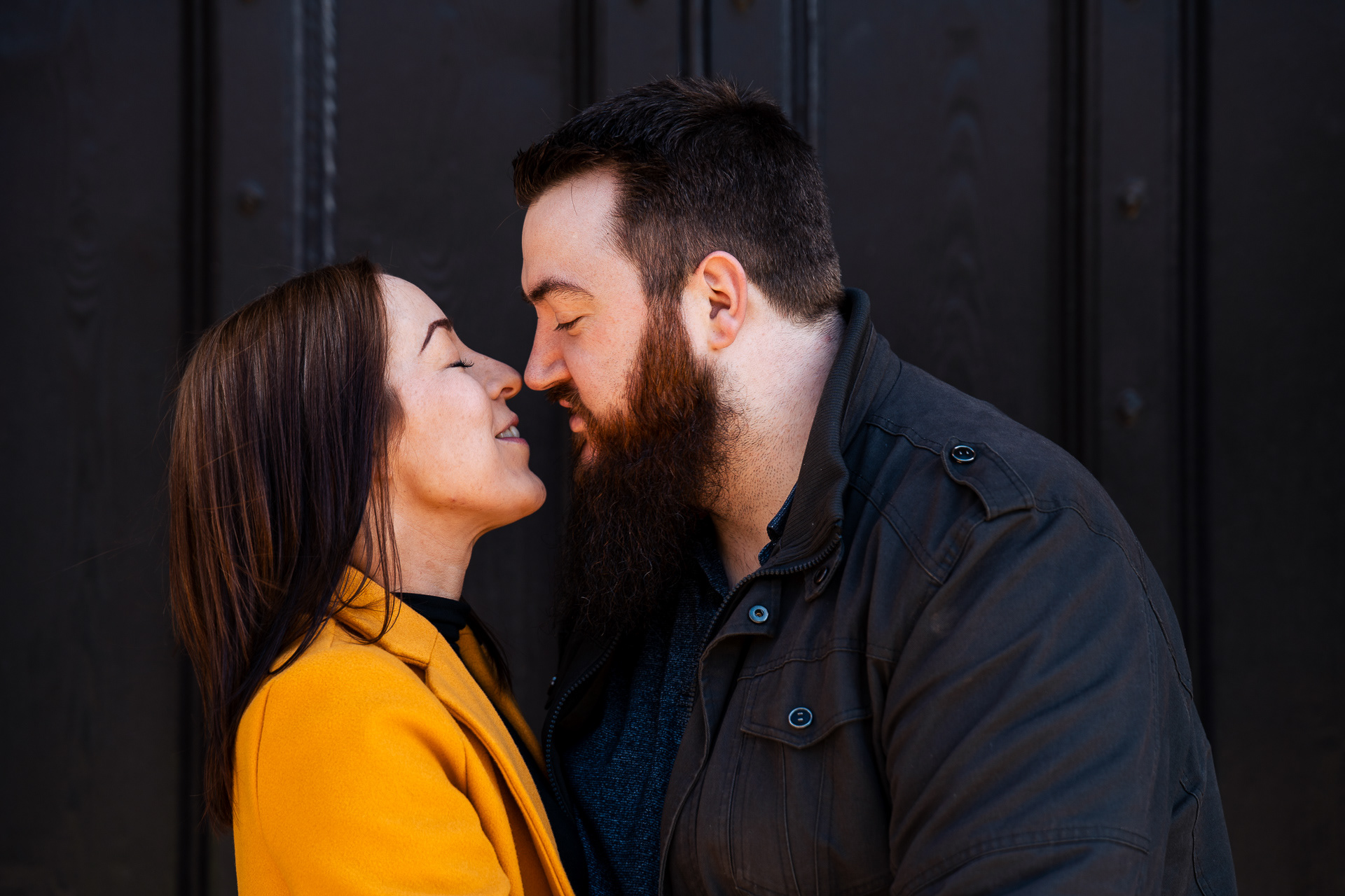 A head and shoulders shot of a couple leaning in, about to kiss. One is a white woman with long dark straight hair and a bright yellow jacket; her partner is a dark haired white man with a bushy beard and a dark jacket. The background is an ornate black door, with details just visible in the light.