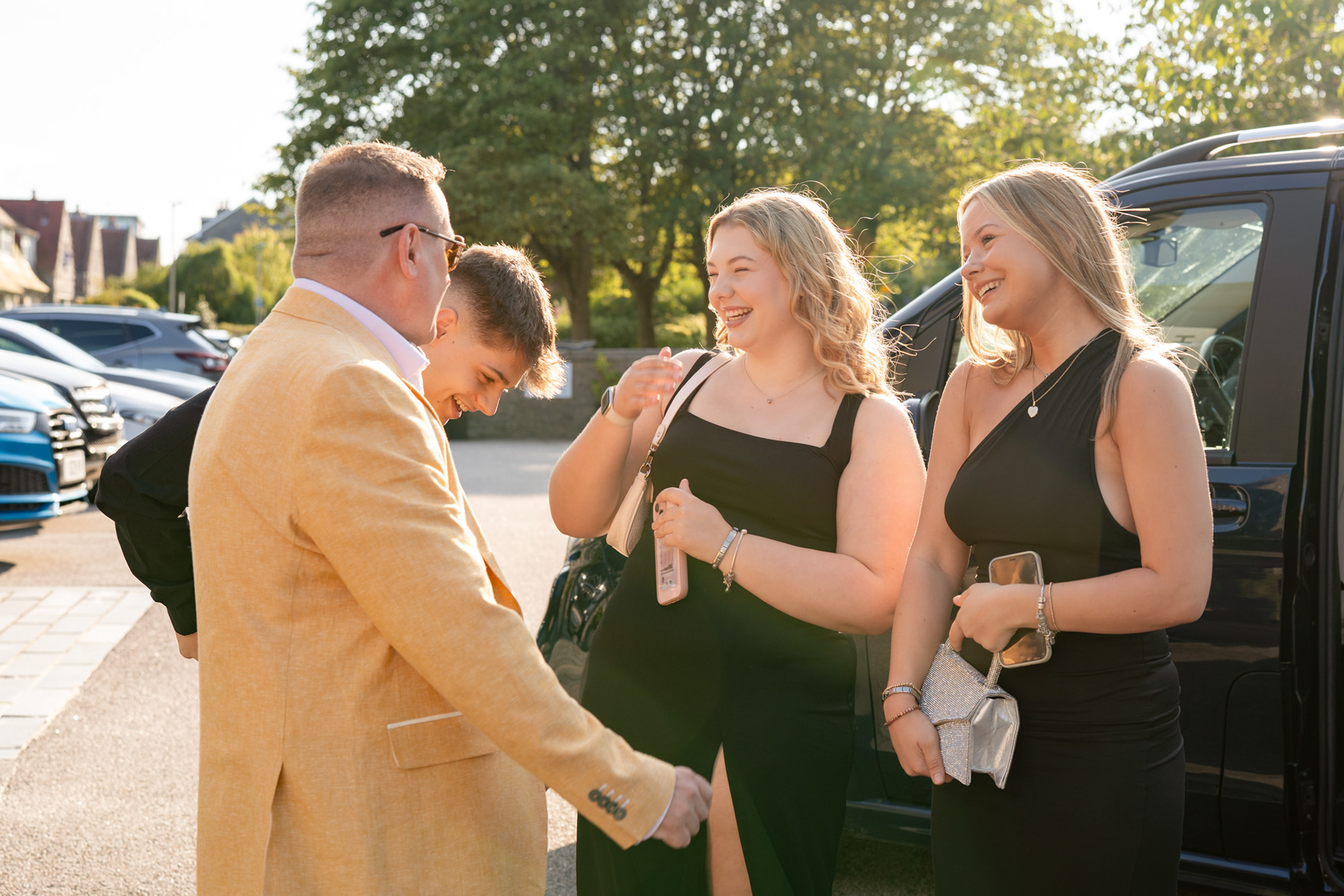 Aberdeen party photography: A man in a yellow suit jacket greets three young adults (two blonde women and a man, all in black) as they step out of a black taxi. They are all lit golden by the sun.