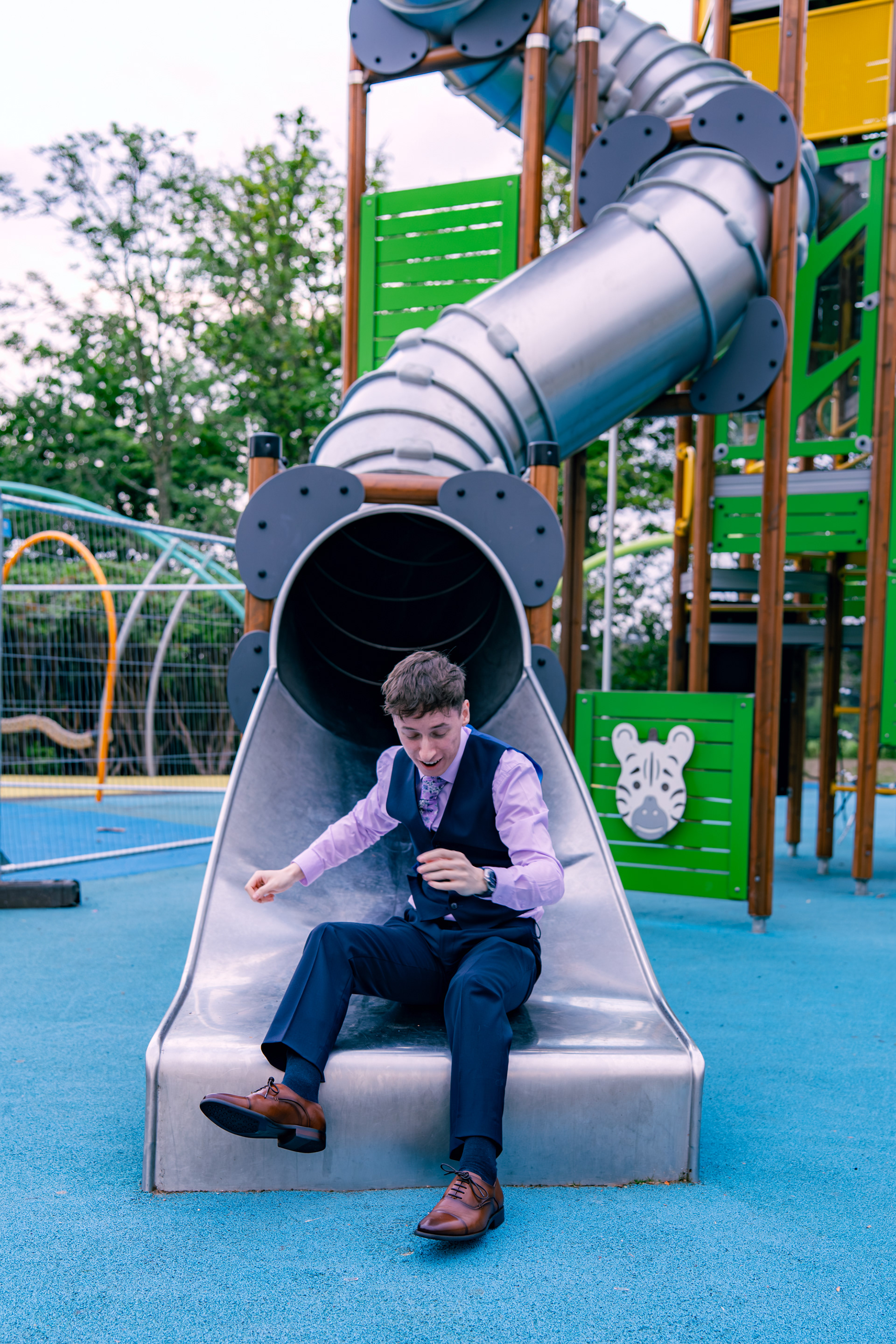 A young man in navy and lilac laughs as he comes down the tube slide during an Aberdeen wedding
