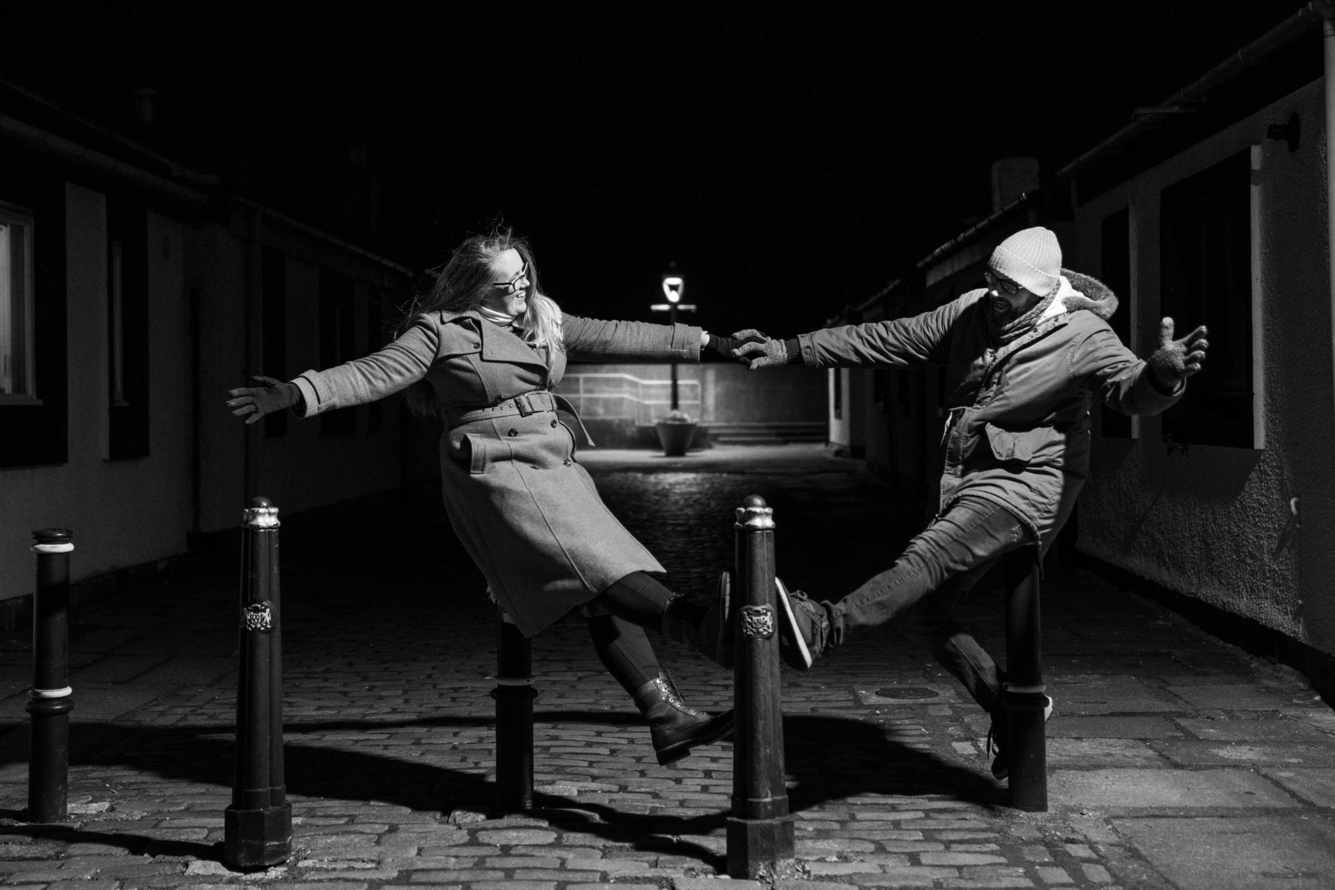 A male-female couple in winter coats balance on top of bollards at Fittie (Footdee) during their Aberdeen couples photo shoot. They are lit by lamplit on a dark night.