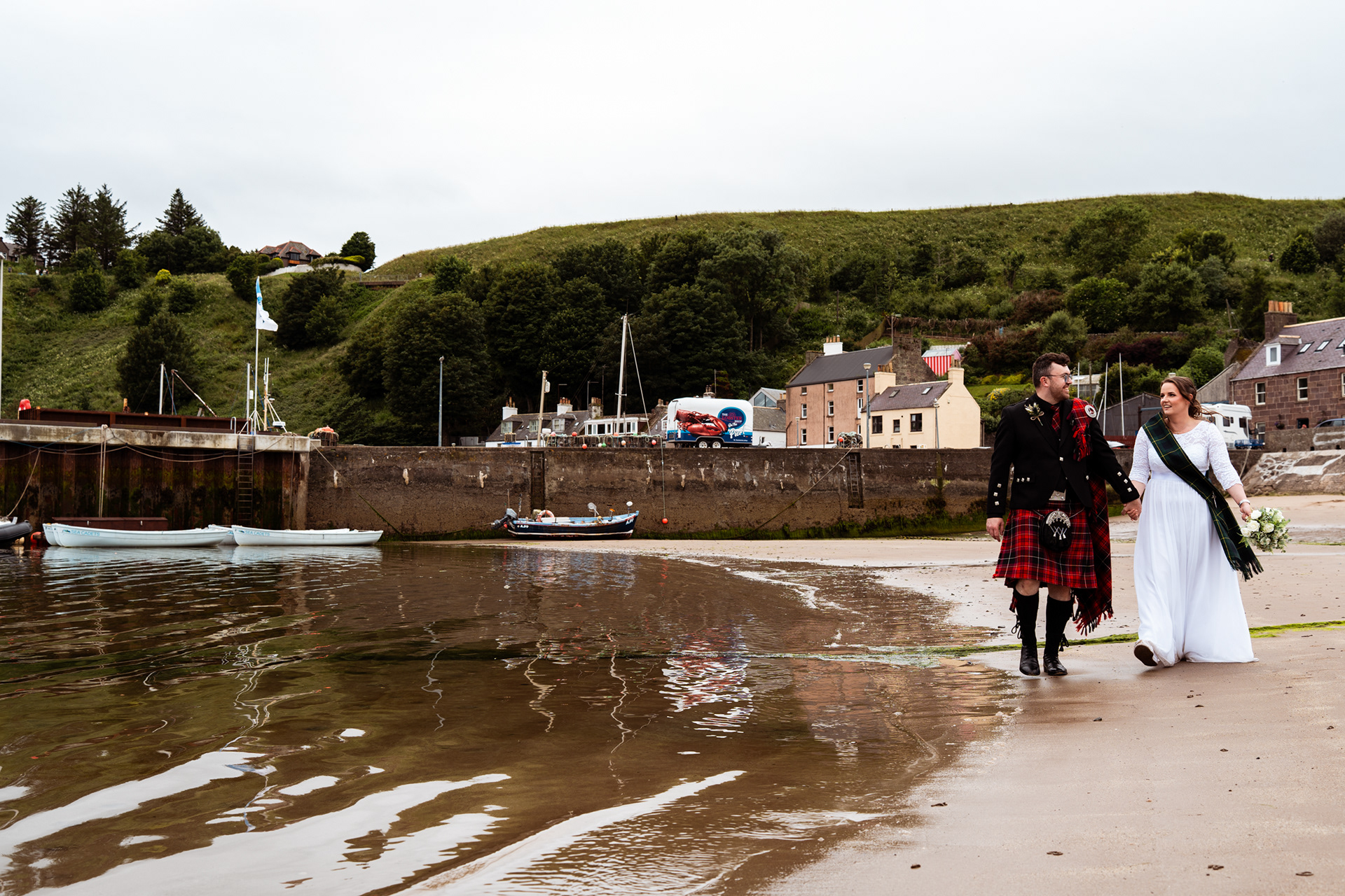 Stonehaven registry office wedding: Bride in white and groom in red kilt walk hand in hand along the sand at Stonehaven Harbour during low tide.