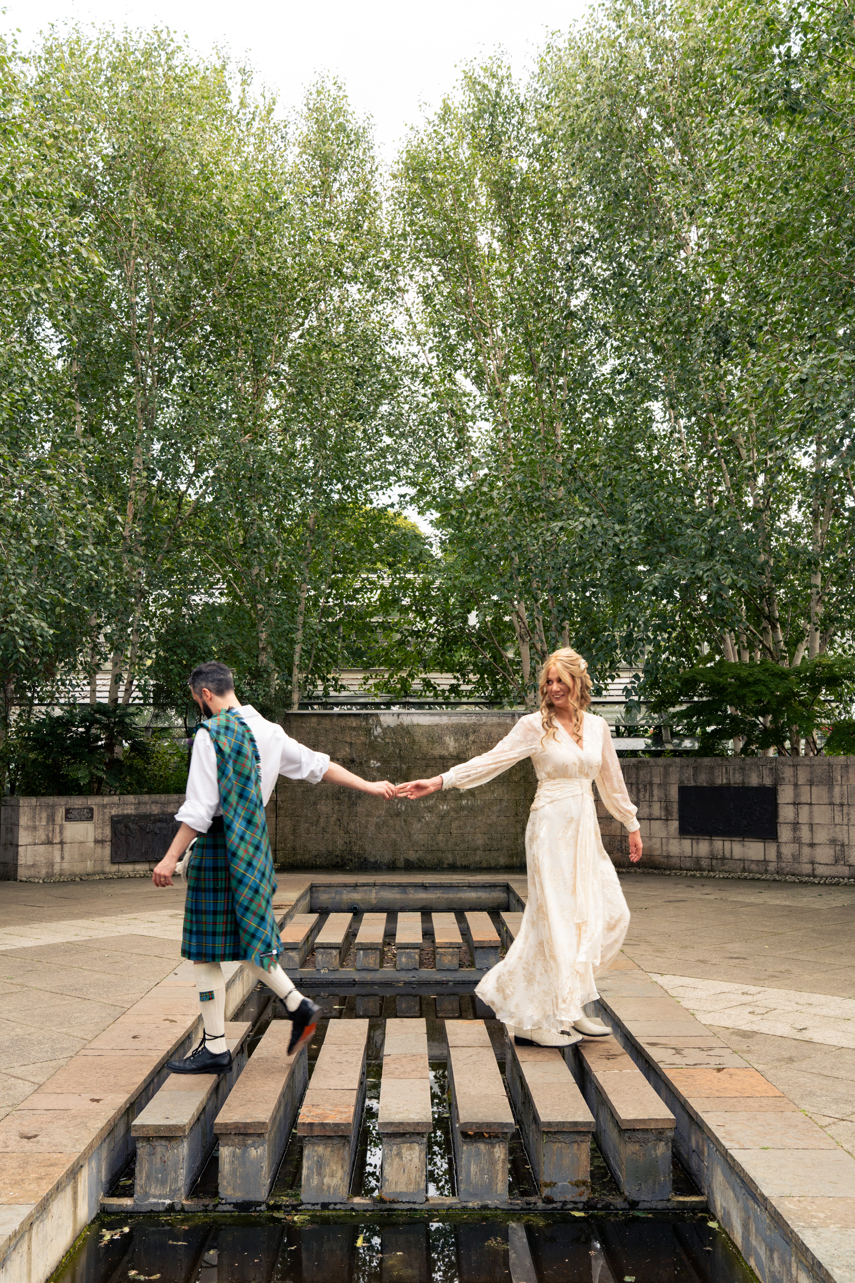 Bride in ivory boho dress and cowboy boots and groom in blue and green kilt walk across the stepping stones at Duthie Park after their Aberdeen Winter Gardens wedding.