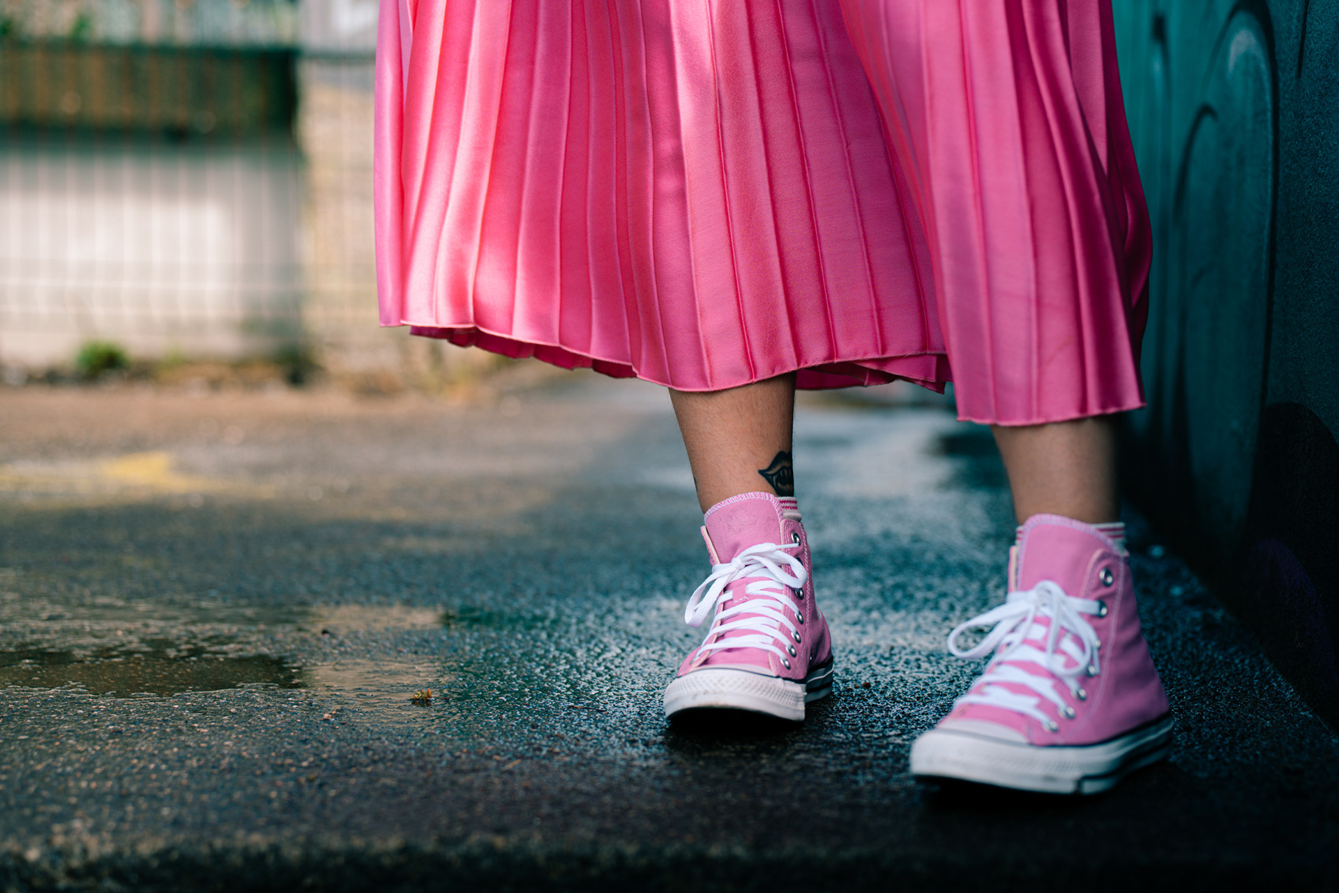 In an urban Aberdeen location, we see a pair of feet in pink Converse, with a pink pleated skirt shining above them. There is a fanged mouth tattooed on one ankle.