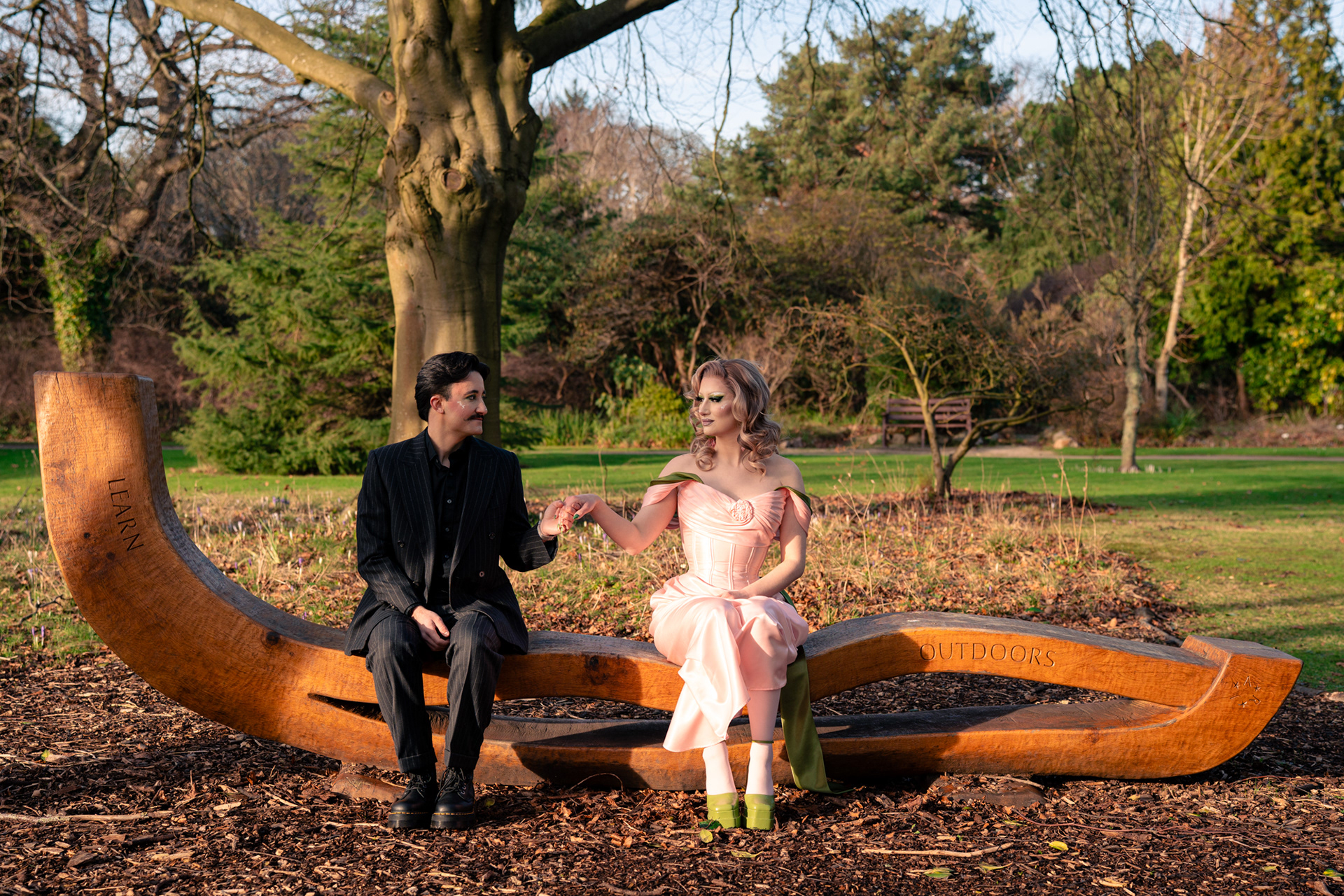 A couple in glamorous drag (they/they and she/her) sit, holding hands, looking at each other on an outdoor bench, lit like a 1920s movie and posed like Wes Anderson.