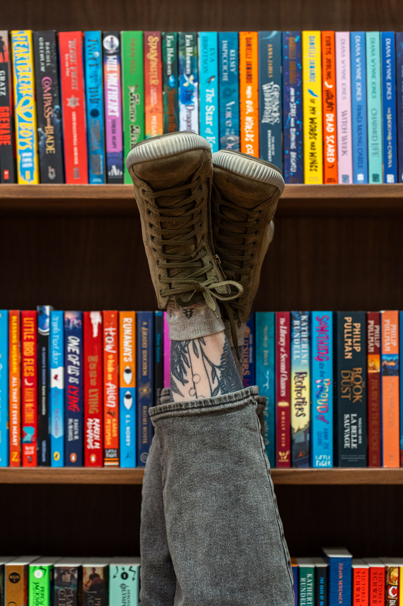 Aberdeen main character shoot: feet in khaki boots and black jeans, with tattoos showing in the gap, are crossed at the ankle, up in the air, leaning against shelves of colourful books.