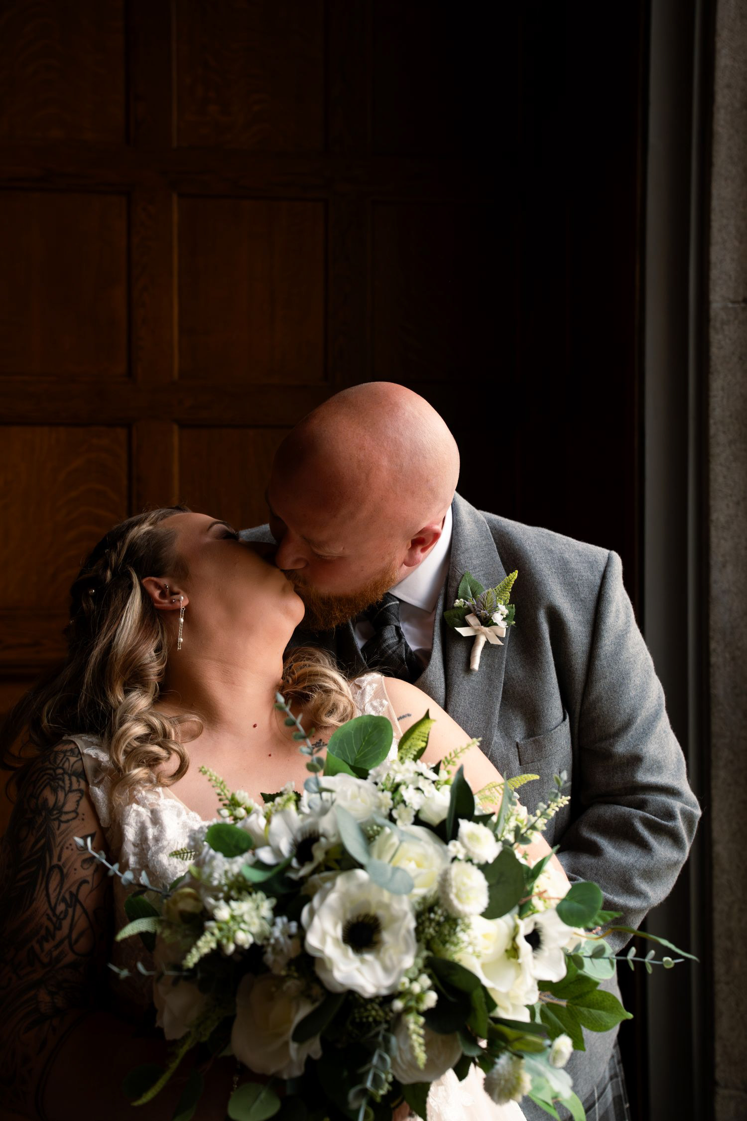 A bride and groom in traditional white dress and kilt outfits, kiss in the Marischal College Grant Room, side lit by window light. Dark panelling shows behind them.