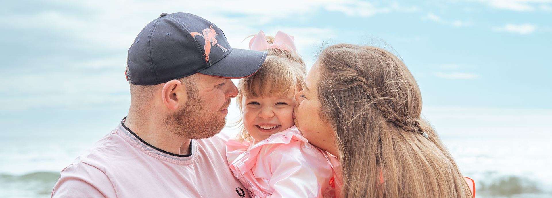 At an Aberdeen family photo shoot, a small girl in pink grins straight at the camera while her mum and dad lean in to kiss her cheeks. The background is blue sky.