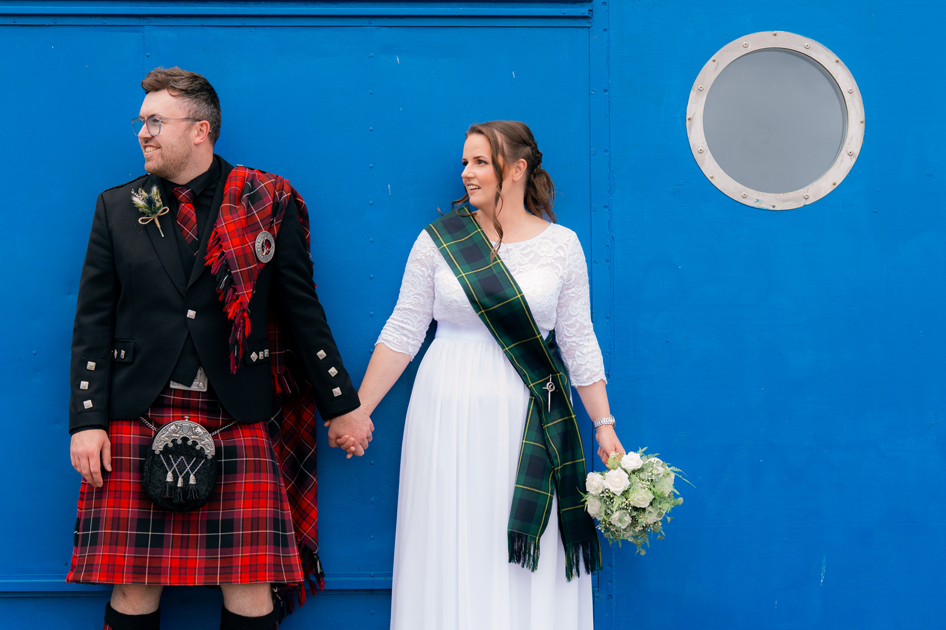 Aberdeenshire wedding: A bride in with and groom in red kilt hold hands against a bright blue background.
