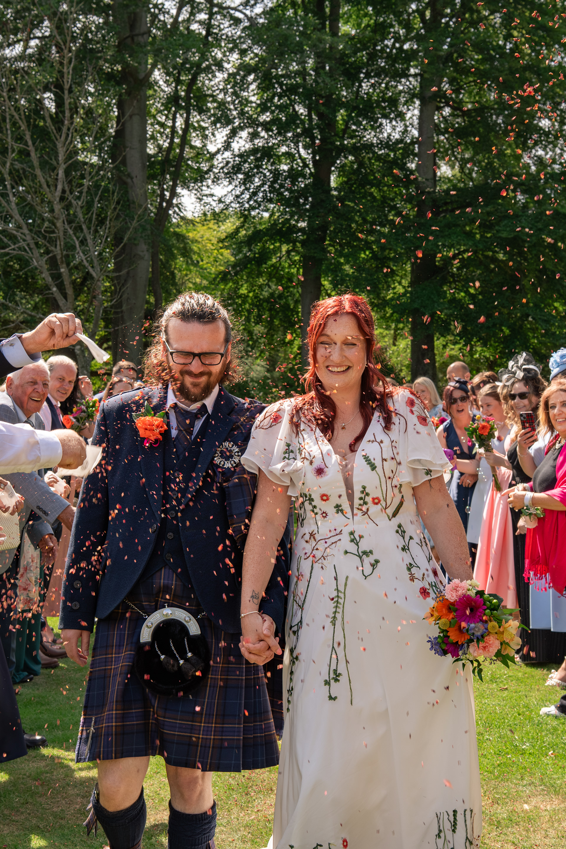 Bride and groom laugh as they are showered with red confetti, walking between their guests on a sunny day in Hazlehead Park