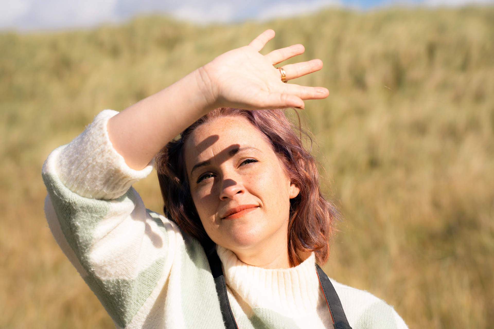 A white woman with ginger and lilac hair and freckles raises her hand above her face to block the sun; the silhouette of her fingers is on her face, her eyes visible in between fingers.