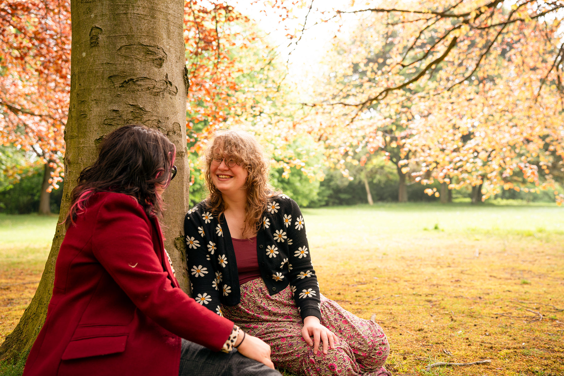 A curly haired woman in a floral cardigan beams at her long-haired boyfriend in red coat, as they sit under autumnal trees with light streaming through them. Aberdeen couple's photography in the park.