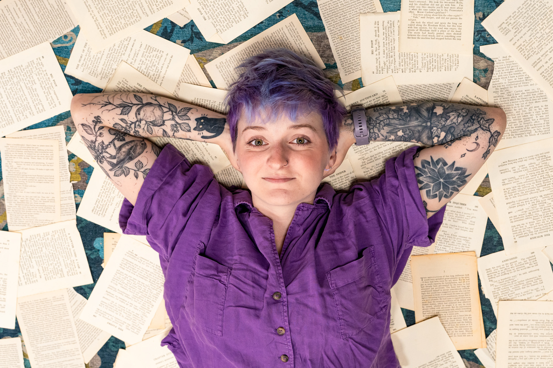 Aberdeen bookshop owner, Christie, a young white woman with purple hair and amazing tattoos, wearing a purple shirt, lies on a floor covered in torn book pages, hands behind her head, looking straight at the camera.