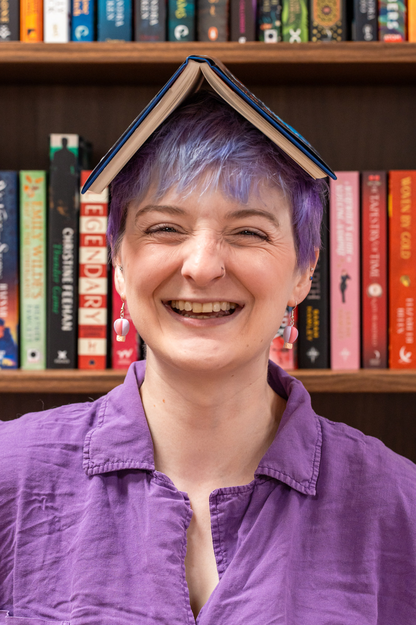 Aberdeen bookshop owner with purple hair and a purple shirt and tiny hot air balloon earrings balances a book on her head and laughs straight at the camera.