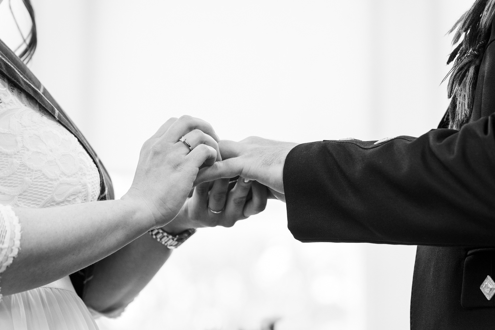 Black and white close up of hands, with bride sliding a ring onto the groom's finger, her own ring visible to the camera.
